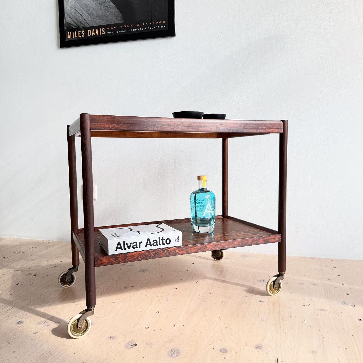 A wooden serving cart with wheels, featuring two shelves. The top shelf displays a blue bottle and a book titled 'Alvar Aalto'. A vintage black-and-white photo of a musician, possibly Miles Davis, is hung on the wall in the background.