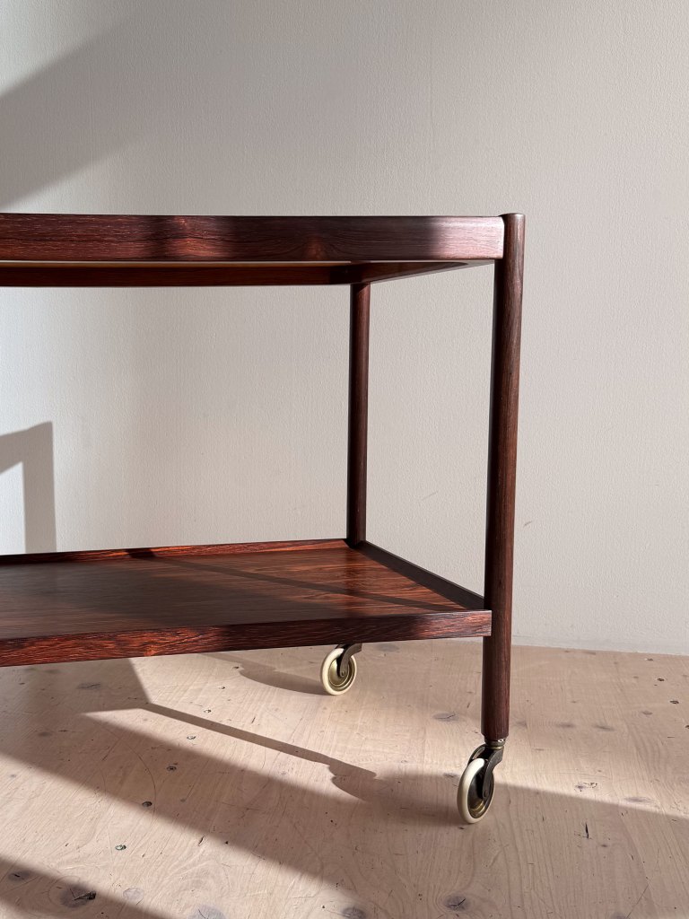 Wooden cart with two shelves and metal wheels, positioned against a light-colored wall.