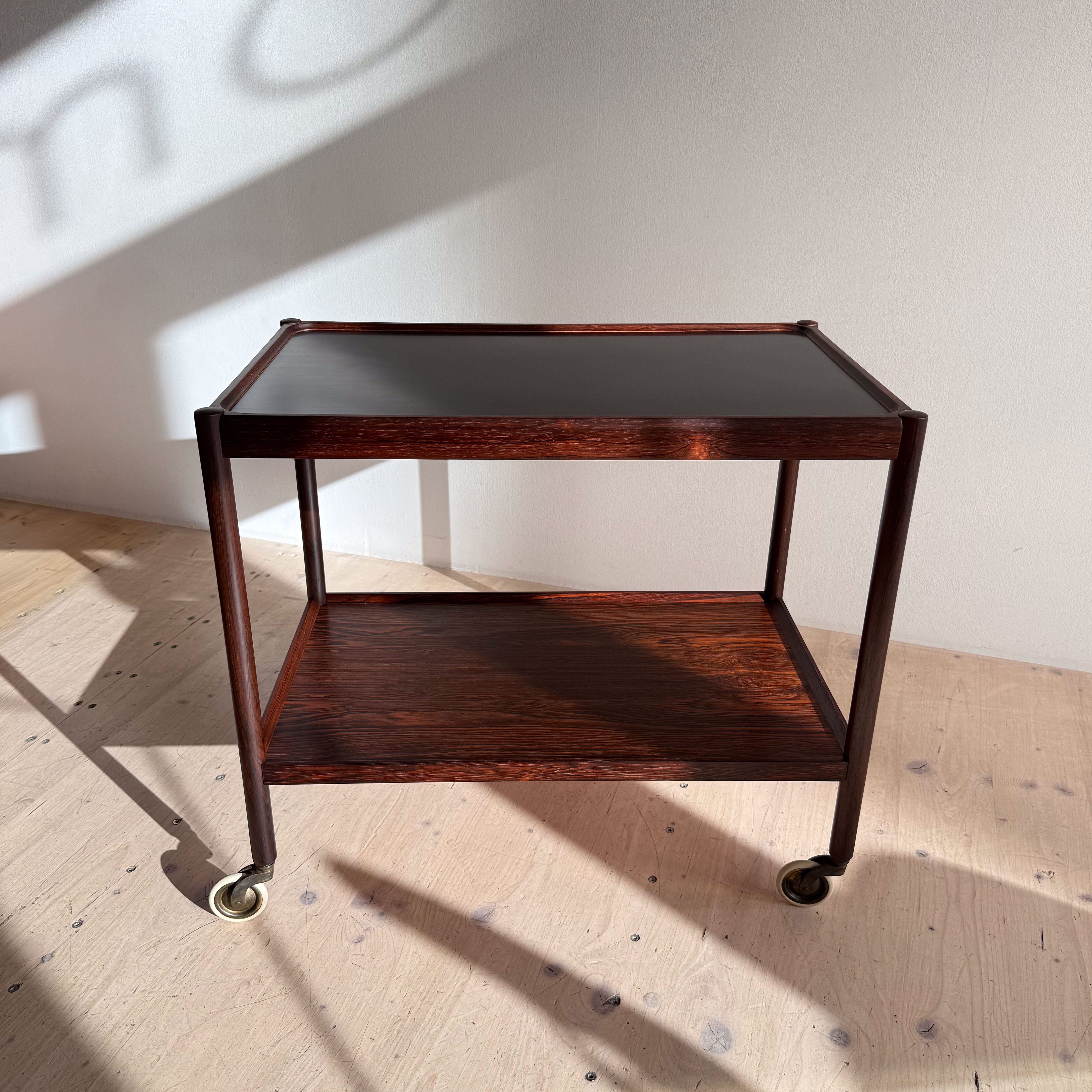A vintage wooden serving cart with a rich finish, featuring two shelves and caster wheels for mobility, set against a softly lit background.