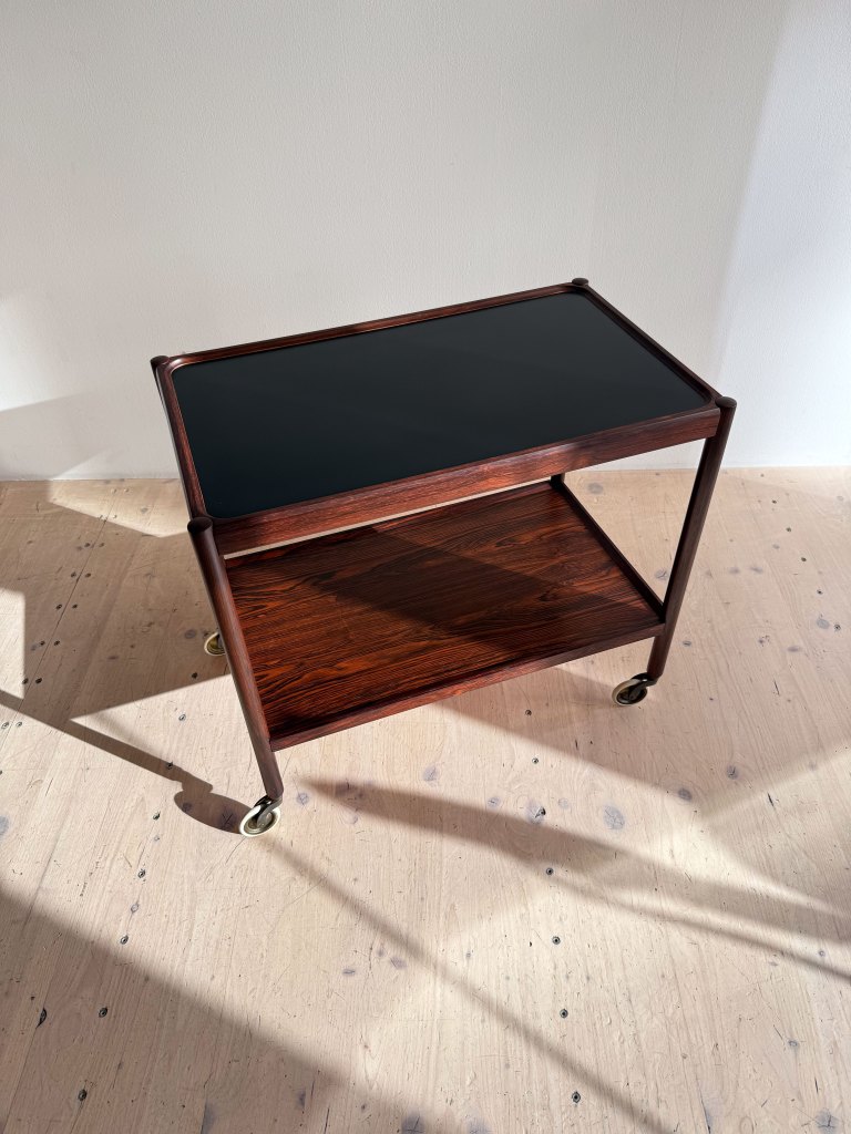 A wooden serving cart with a black top and a lower shelf, featuring circular wheels for mobility.