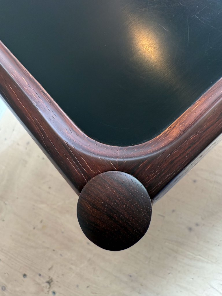 Close-up view of the corner of a wooden table with a circular leg and a glossy black surface.