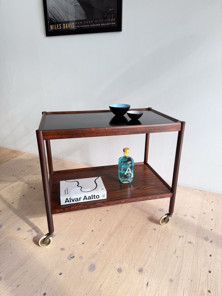 A wooden serving cart with two tiers, featuring a black tabletop. On the top shelf, there are two bowls, one blue and one white, alongside a book titled 'Alvar Aalto'. The bottom shelf holds a bottle of teal-colored liquor. A framed poster of Miles Davis hangs on the wall behind.