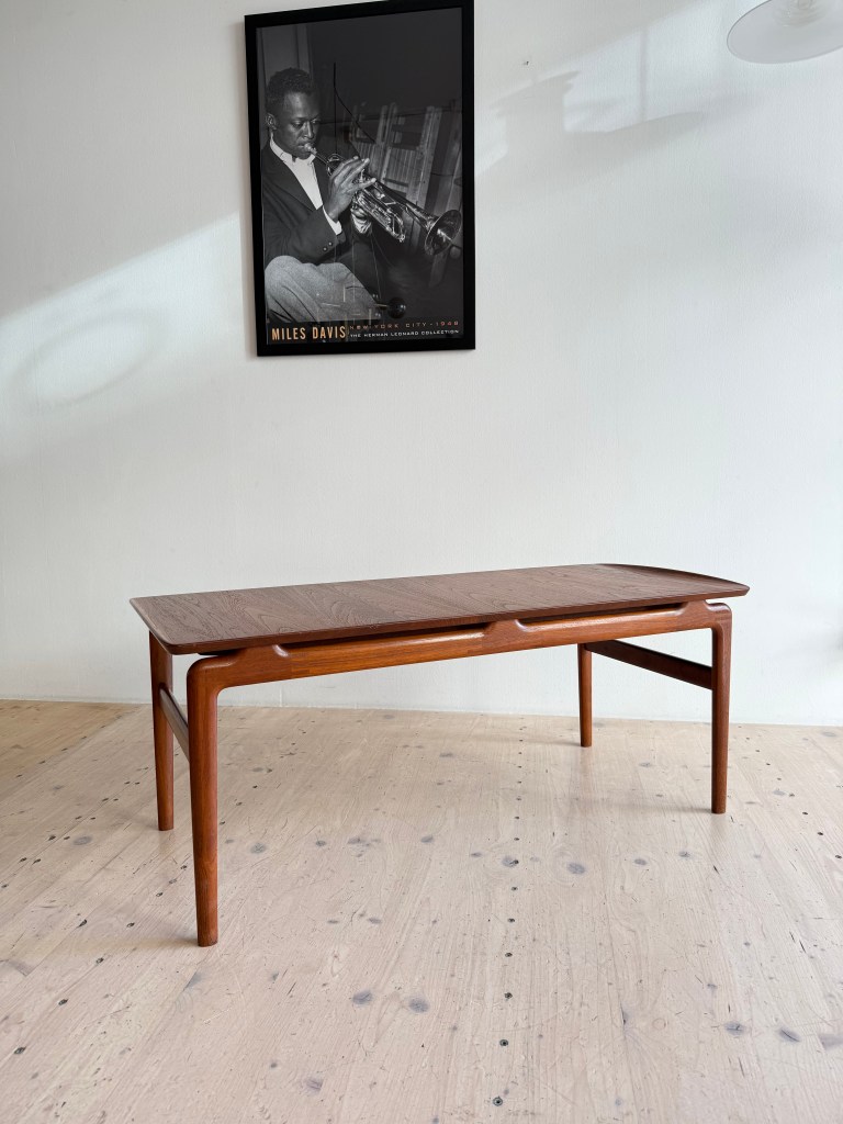 A wooden coffee table with a sleek design, positioned on a light wooden floor, accompanied by a vintage black and white poster of a musician playing the trumpet.