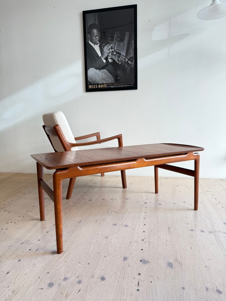 A modern wooden coffee table with a unique design sits on a light wooden floor, accompanied by a stylish armchair, against a wall featuring a framed black-and-white poster of a musician playing the trumpet.