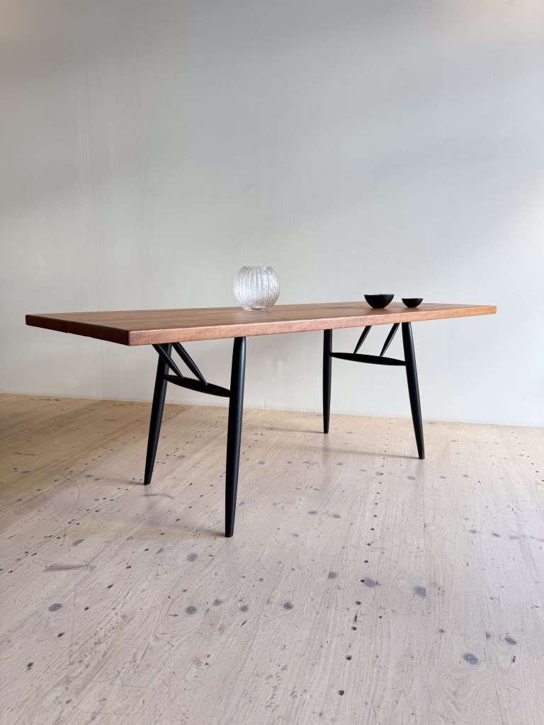 A modern wooden table with a minimalist design, featuring black legs and a polished surface. On top, there is a glass vase and two small bowls, set against a neutral wall and wooden floor.