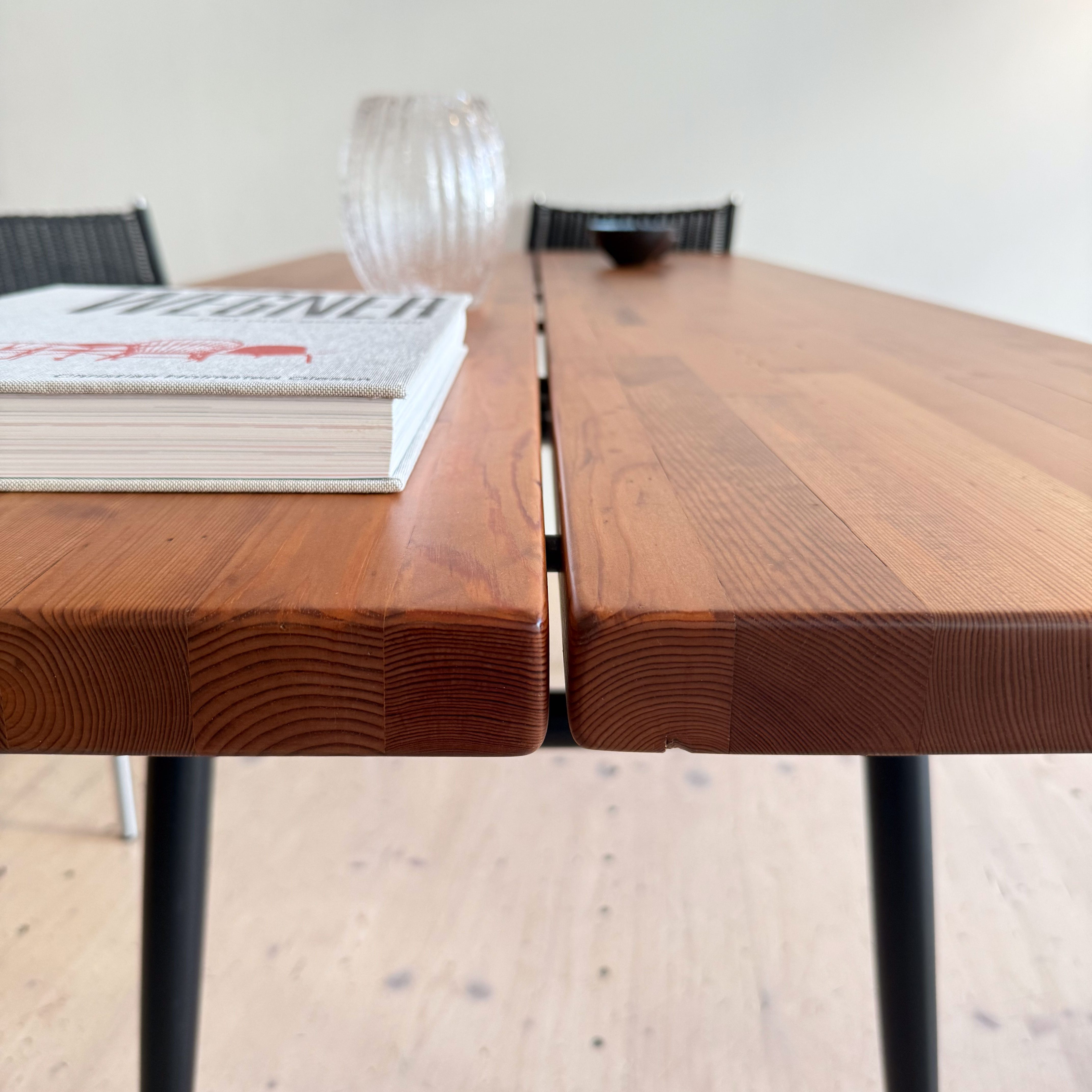 A close-up view of a wooden dining table with a smooth surface, featuring a book and a decorative glass vase on it, set against a neutral wall background.