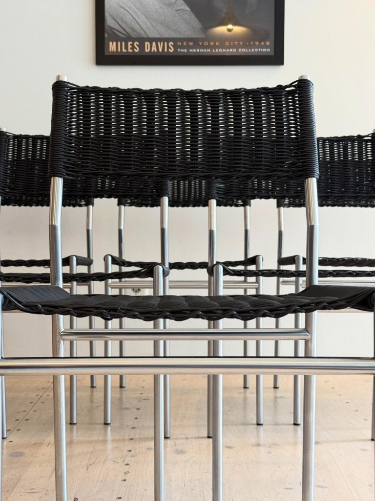 A row of modern black woven chairs with metallic frames, viewed from the front, showcasing a minimalist design against a light wooden floor.