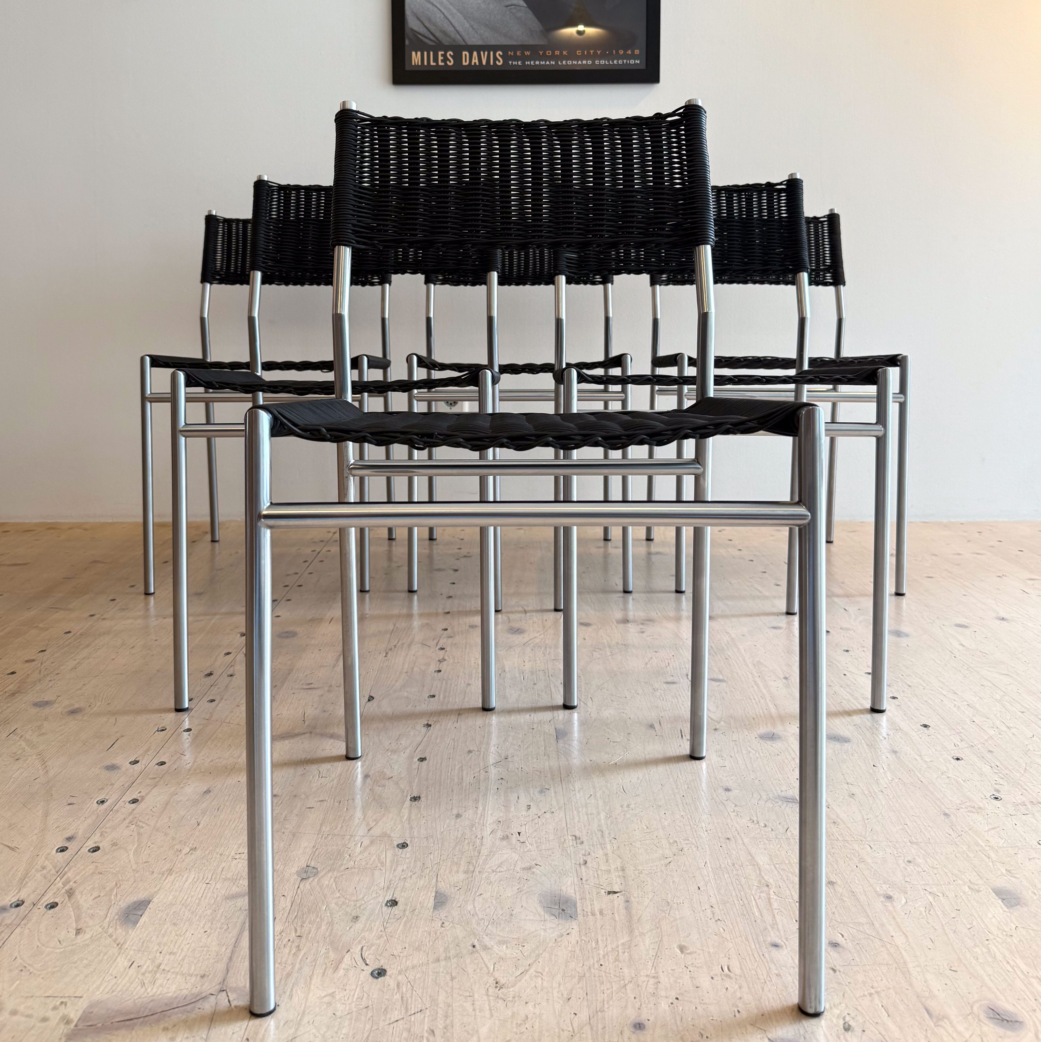 A set of black woven dining chairs with metal legs, arranged in front of a white wall featuring a framed black and white photograph of a musician playing the trumpet.