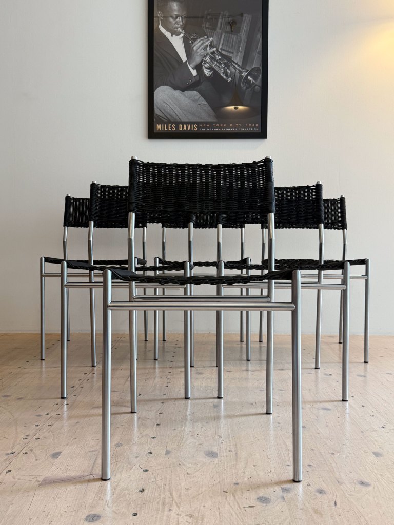 A row of black woven chairs with silver metal frames positioned in front of a framed black and white poster of Miles Davis.