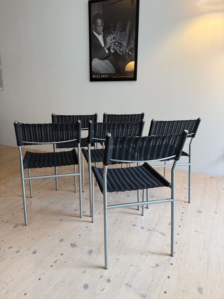 A set of five modern black chairs arranged in a room with a wooden floor, featuring a poster of a jazz musician on the wall.