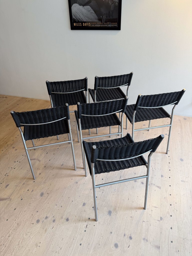 A set of six black woven chairs arranged in a circle on a wooden floor, with a poster of Miles Davis visible on the wall in the background.