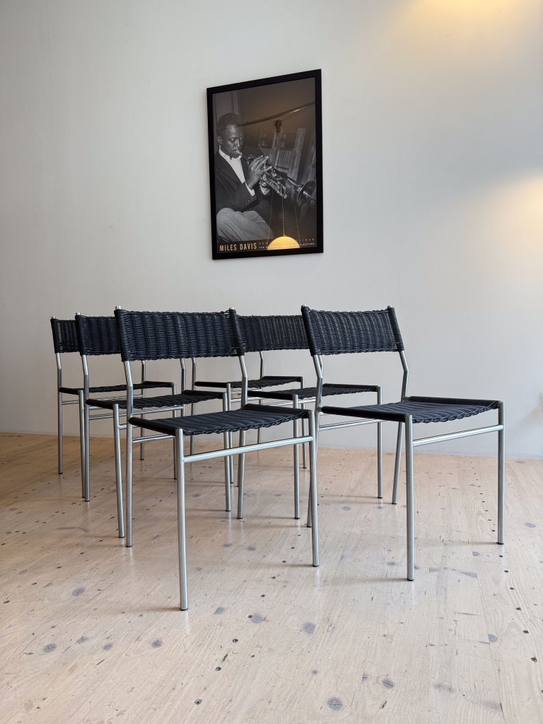 A modern setting with six black woven chairs arranged in front of a wall featuring a framed black and white poster of a jazz musician.