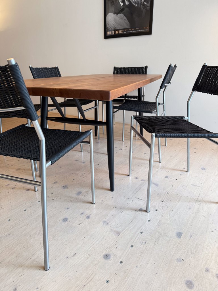 A wooden dining table with black woven chairs in a minimalist setting, featuring light wooden flooring and a framed poster on the wall.