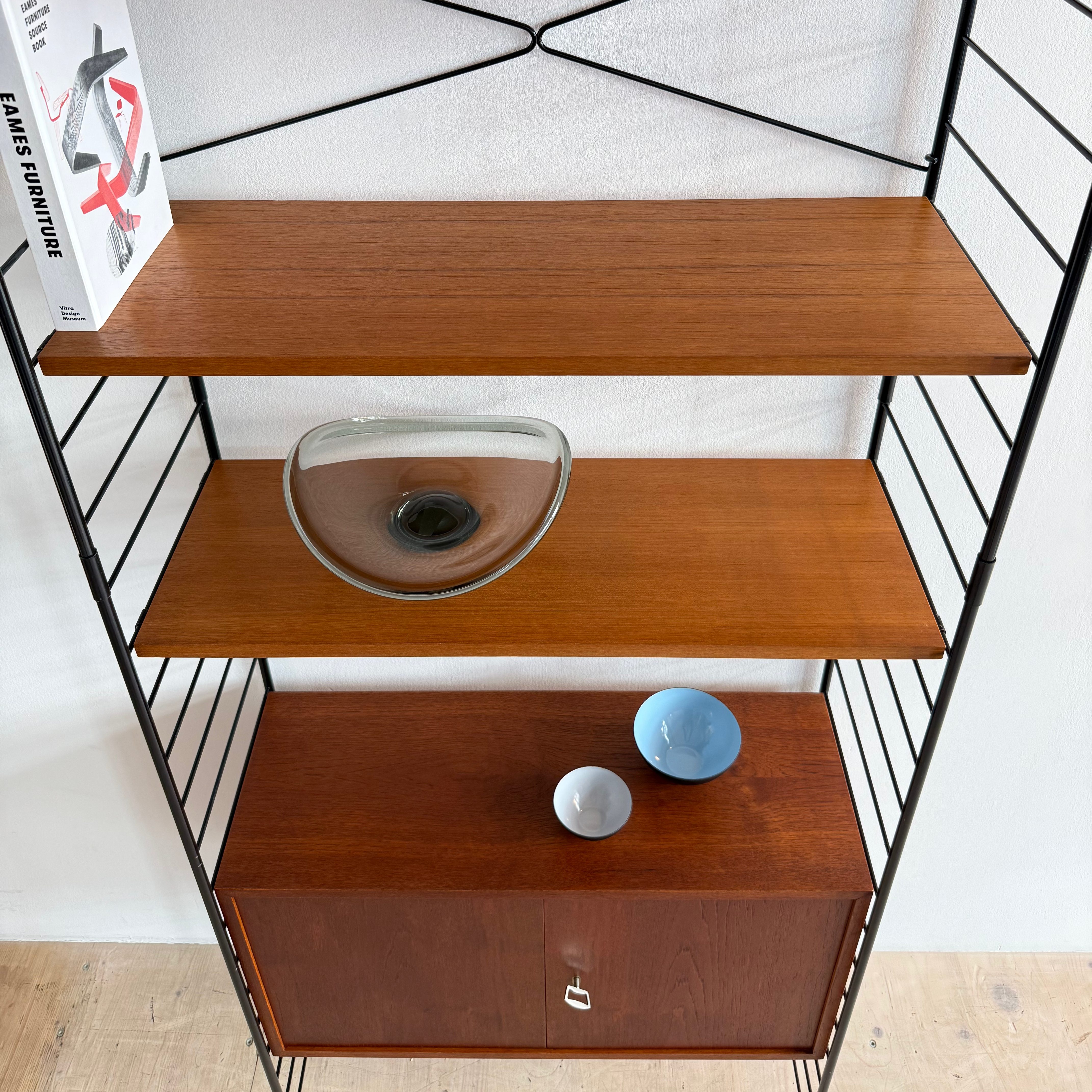 A modern shelving unit with wooden shelves, featuring a decorative glass bowl, two small bowls, and a book titled 'Modern Furniture Design.' The shelf has a sleek black metal frame.