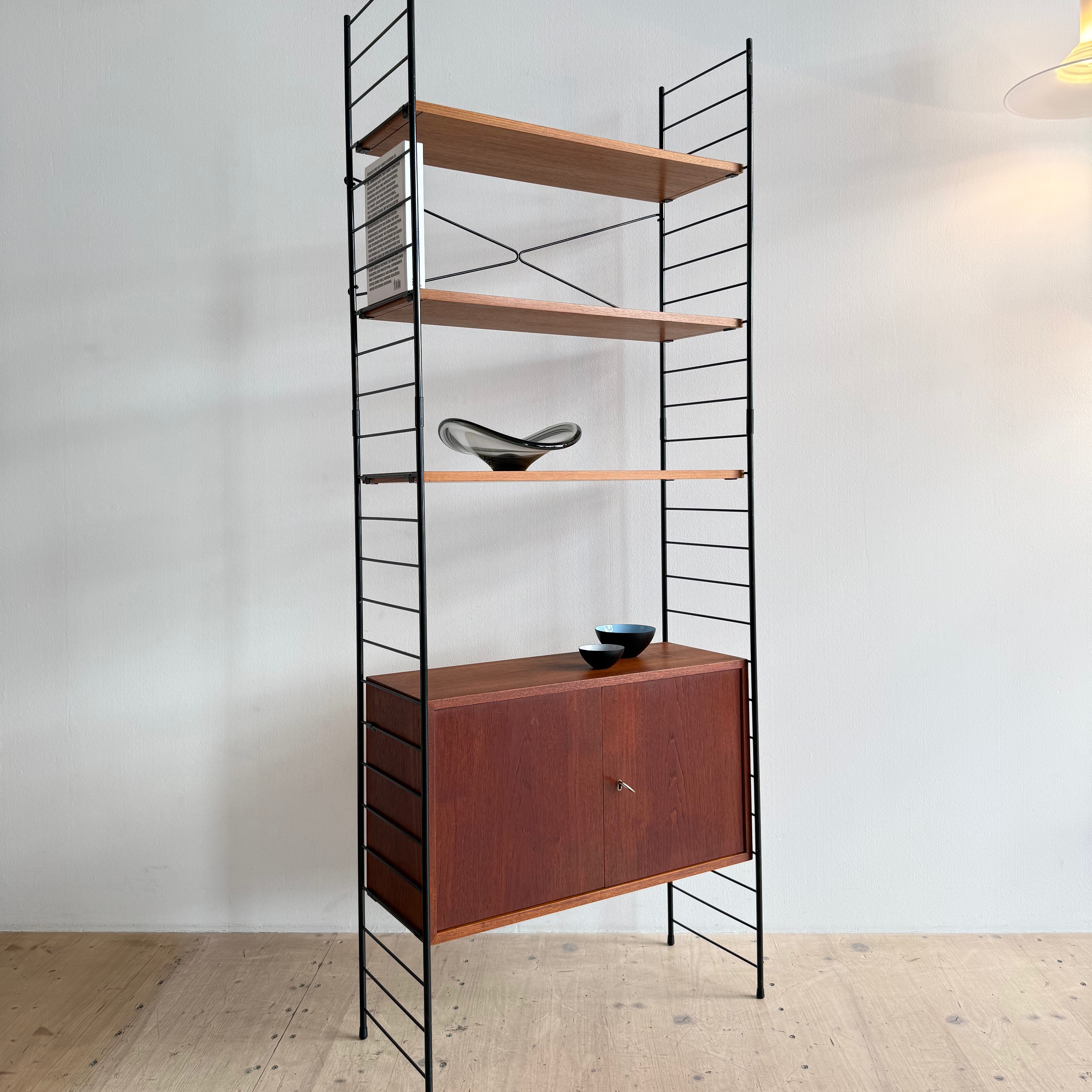 A modern wooden shelving unit with metal accents, featuring three open shelves and a lower cabinet. Decorative objects sit on the shelves, against a light-colored wall.