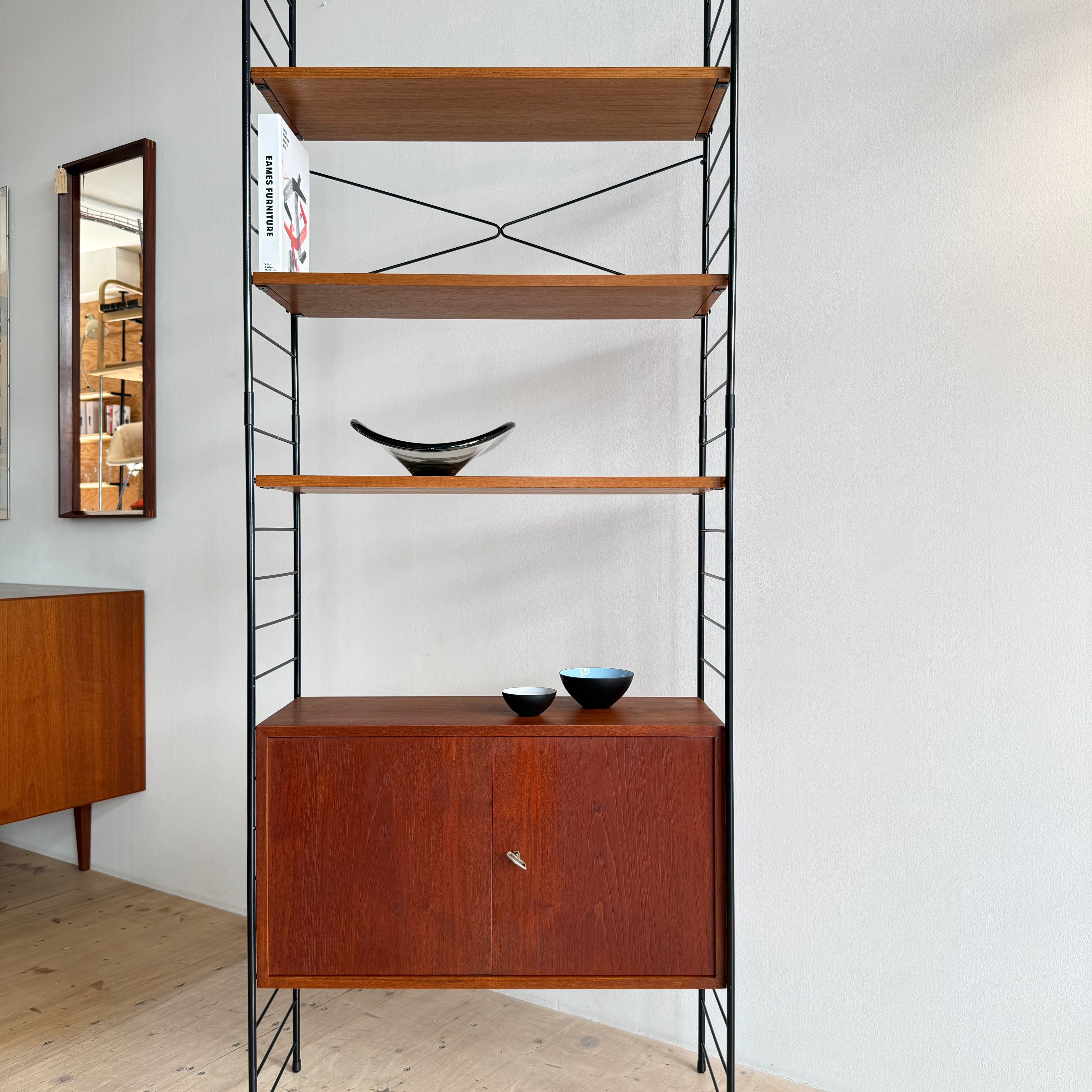 A modern wooden shelving unit with black metal supports, featuring three open shelves and a lower cabinet. The shelves hold decorative items, including a sleek black bowl.