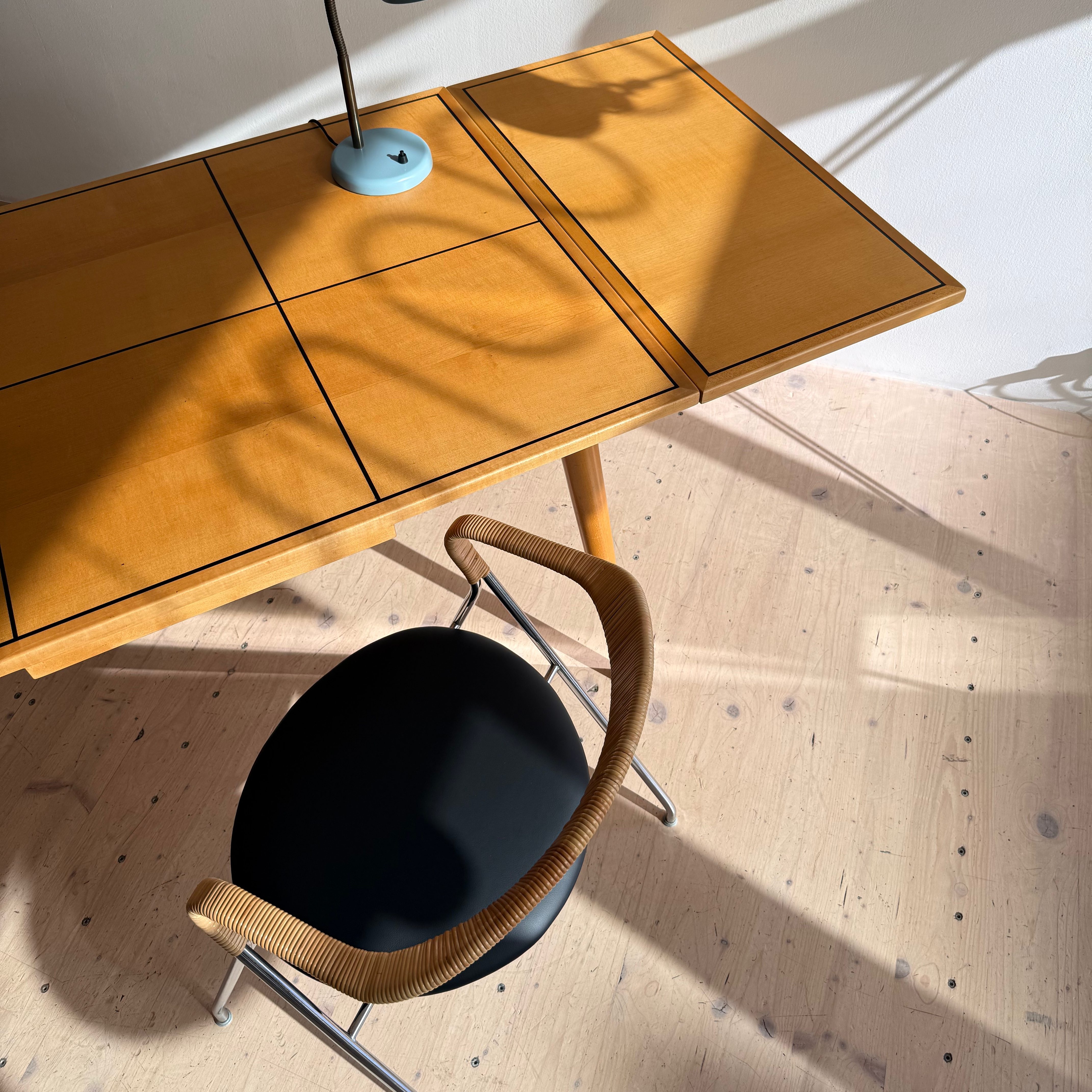 A wooden desk with a blue lamp casting shadows, accompanied by a stylish black chair with a woven armrest.