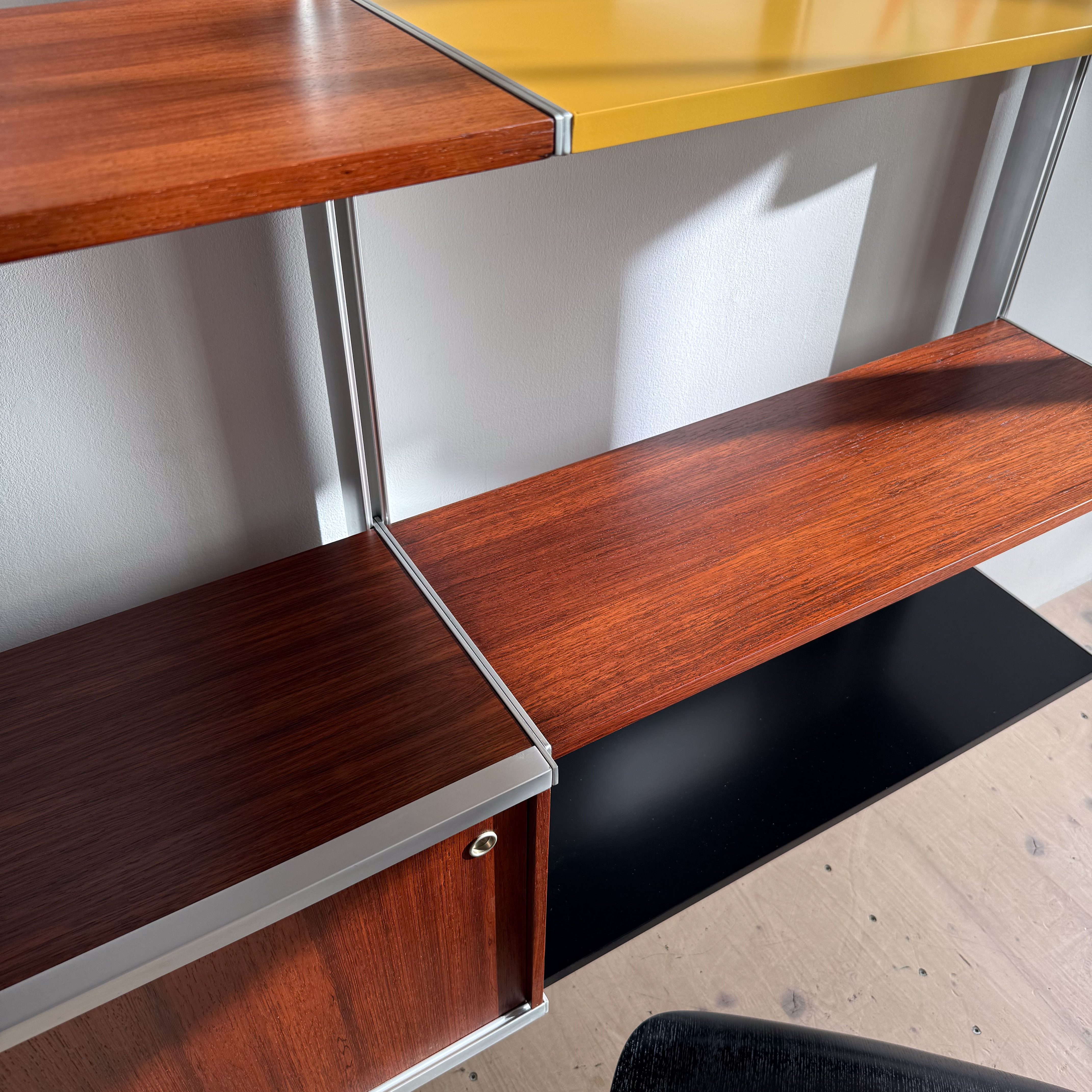 Close-up of a modern shelving unit featuring wooden shelves in various finishes, including a yellow top shelf, a brown middle shelf, and a black bottom shelf, with a decorative book displayed on the top.