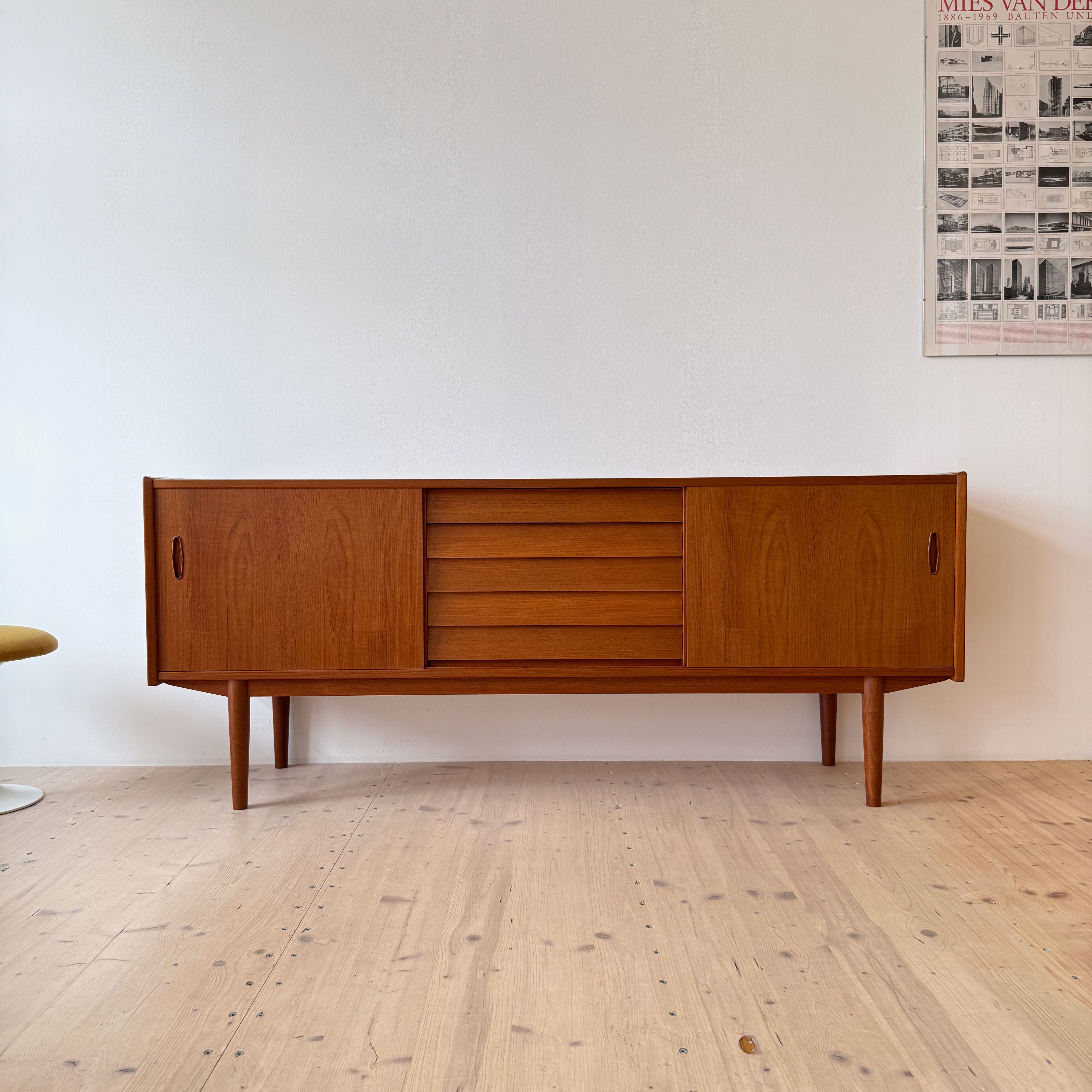 A wooden mid-century modern sideboard with closed cabinets and horizontal slits, set against a plain wall with a poster in the background.