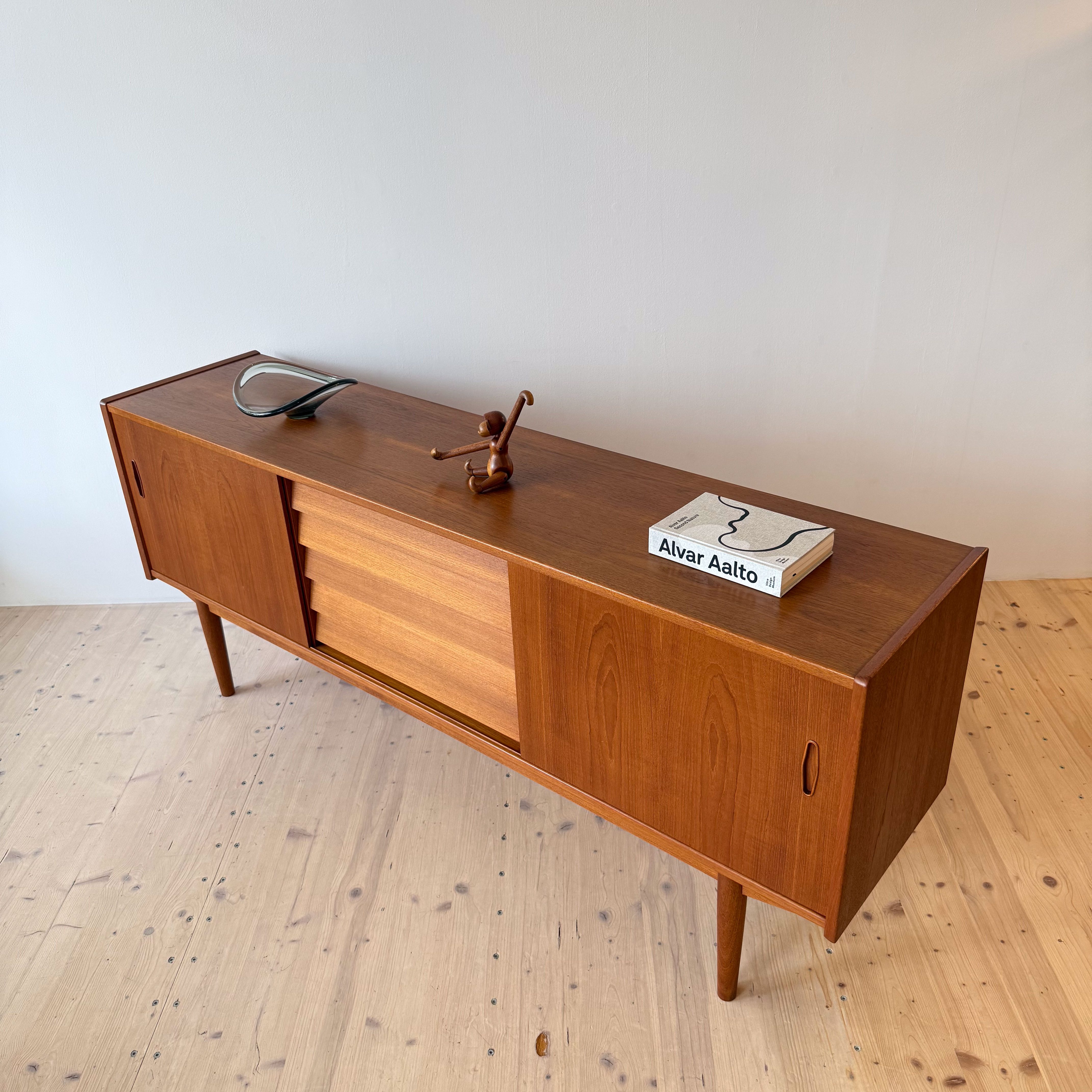 A mid-century modern wooden sideboard with a minimalist design, featuring sliding doors and tapered legs, topped with a decorative sculpture and a book by Alvar Aalto.