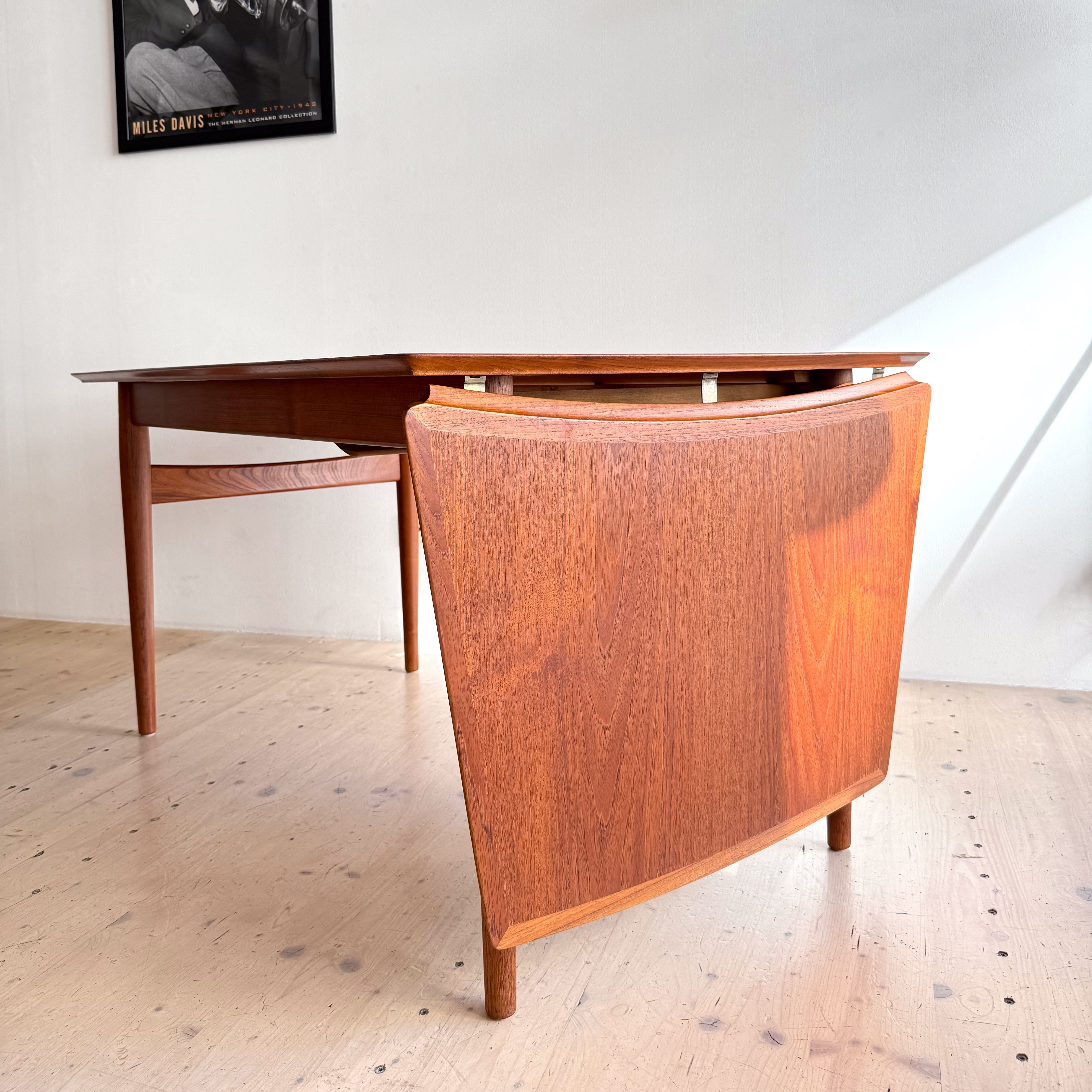 A wooden dining table with a curved chair in front, positioned against a white wall. A black and white poster of a musician is visible in the background.