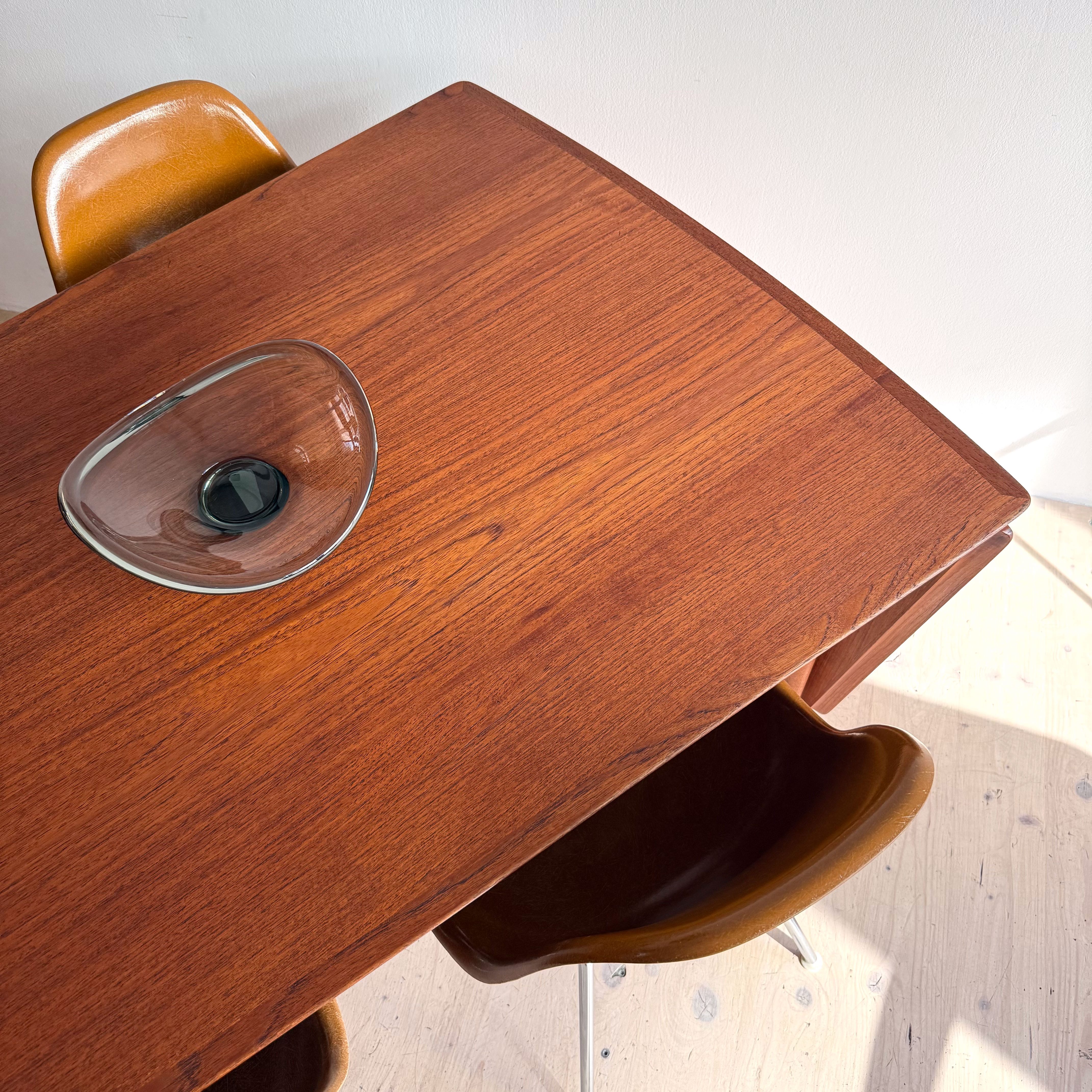 A wooden dining table with a sleek surface, accompanied by two mid-century modern chairs. A glass bowl is centered on the table, reflecting light in a bright, minimalist setting.
