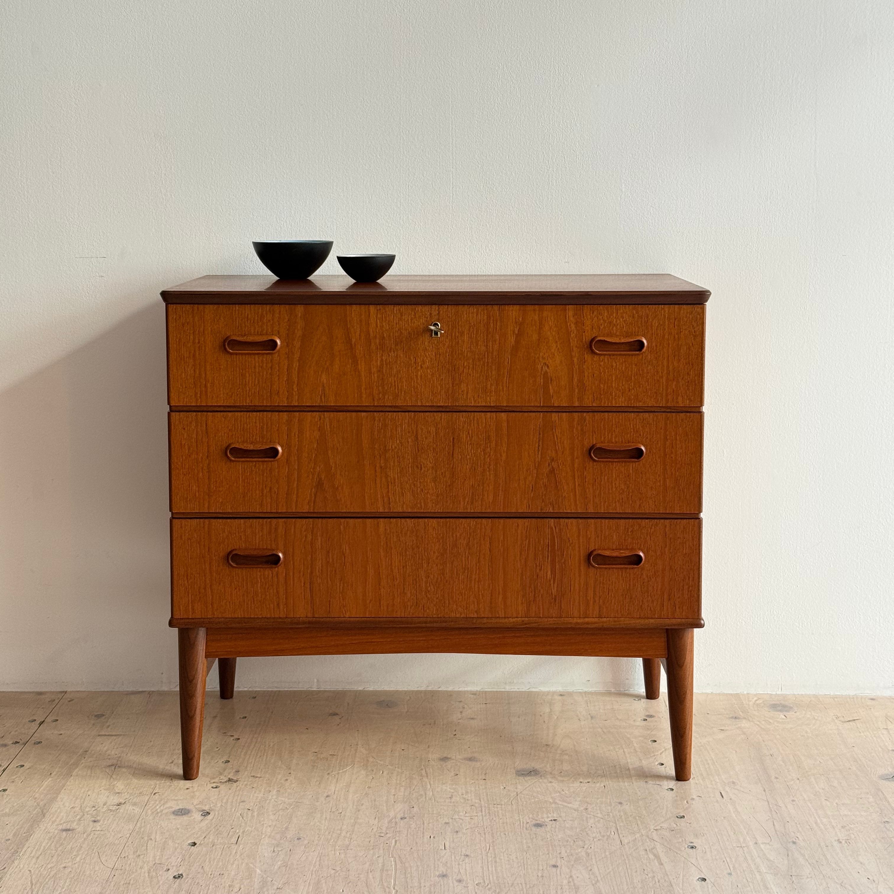 A mid-century modern wooden dresser with three drawers and tapered legs, placed against a white wall, accompanied by two small black bowls on top.