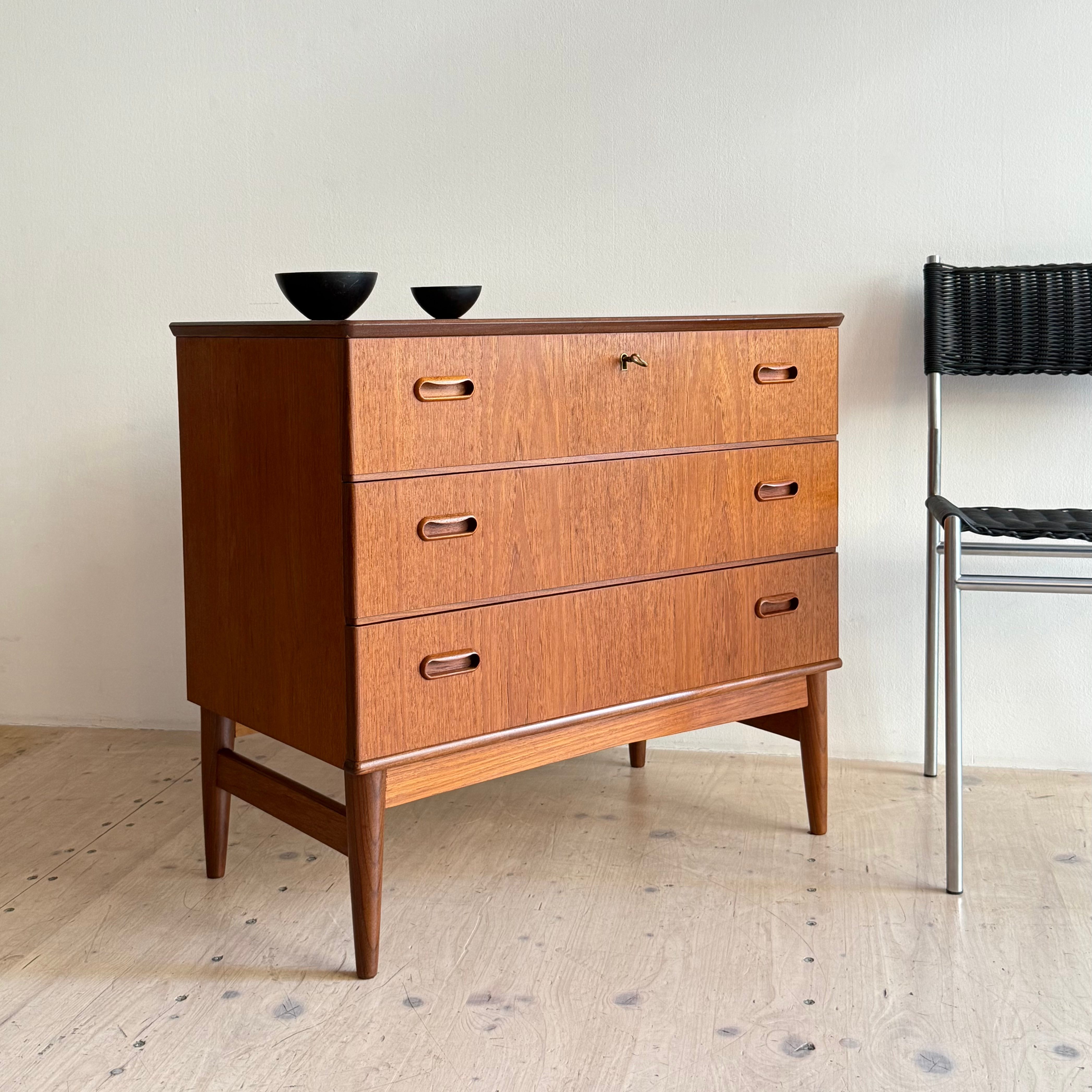 A mid-century wooden dresser featuring three drawers, round handles, and tapered legs, accompanied by two small black bowls and a modern chair in a minimalist setting.