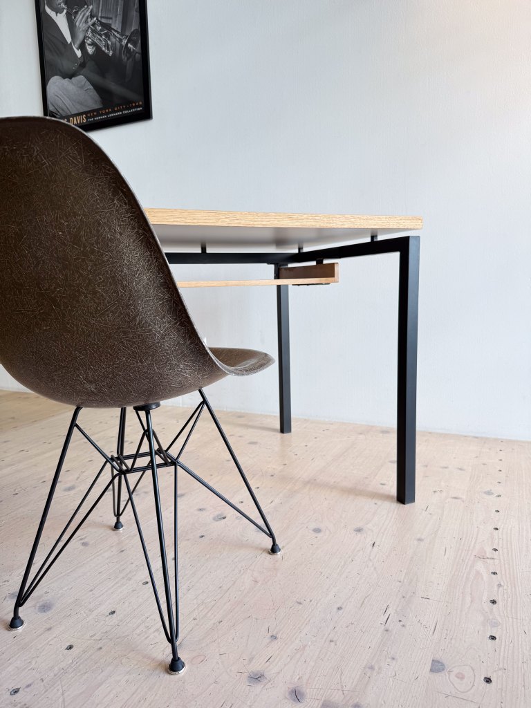 A modern workspace featuring a sleek wooden desk with a black metal frame and a stylish brown chair with an open design. The chair's legs are made of thin black wires, and a vintage poster is visible on the wall behind.