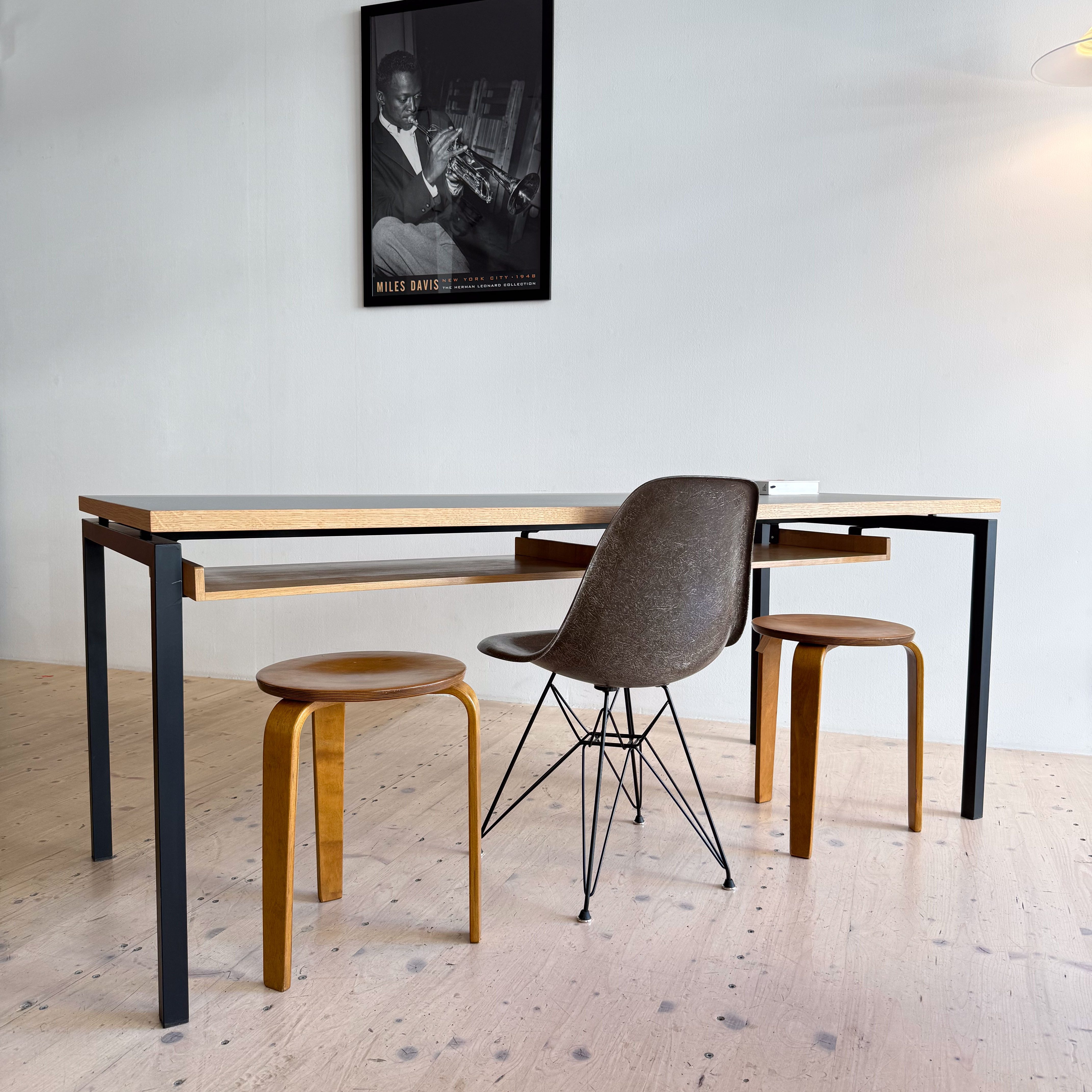 A modern dining area featuring a minimalist wooden table with black legs, two wooden stools, and a stylish chair with a wire base. A poster of Miles Davis hangs on the wall, adding a touch of artistic flair to the space.