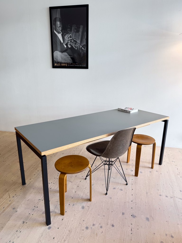 A minimalist dining area featuring a long gray table with wooden edges, accompanied by three diverse chairs: a metal base chair, a round wooden stool, and a modern design chair. A black and white framed poster of a musician hangs on the wall in the background.