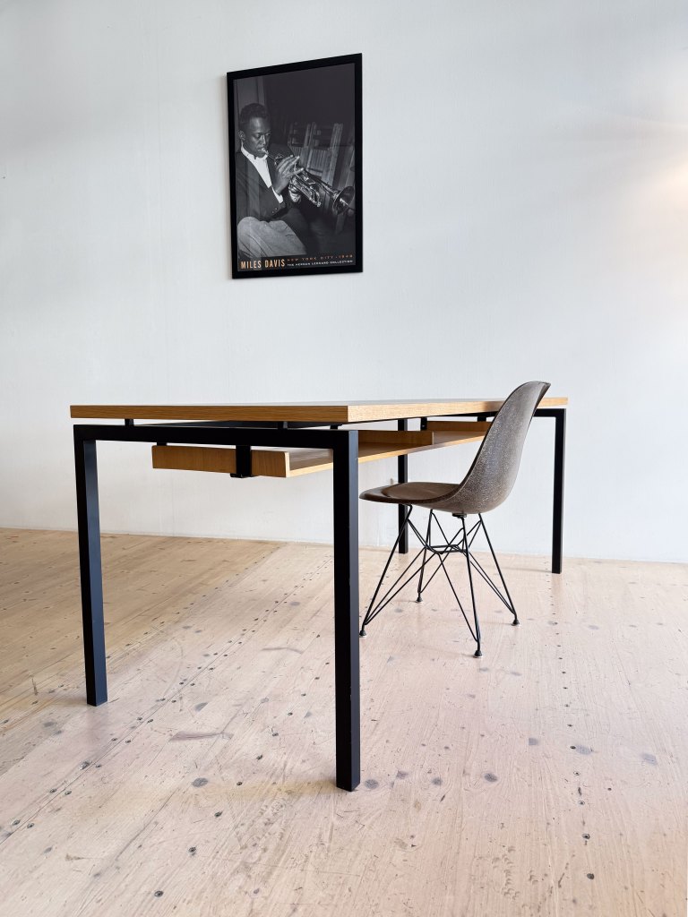 A modern desk with a wooden top and black legs, featuring a single drawer. Beside it is a stylish chair with a soft fabric seat and metal legs. A black and white poster of a musician hangs on the wall above.