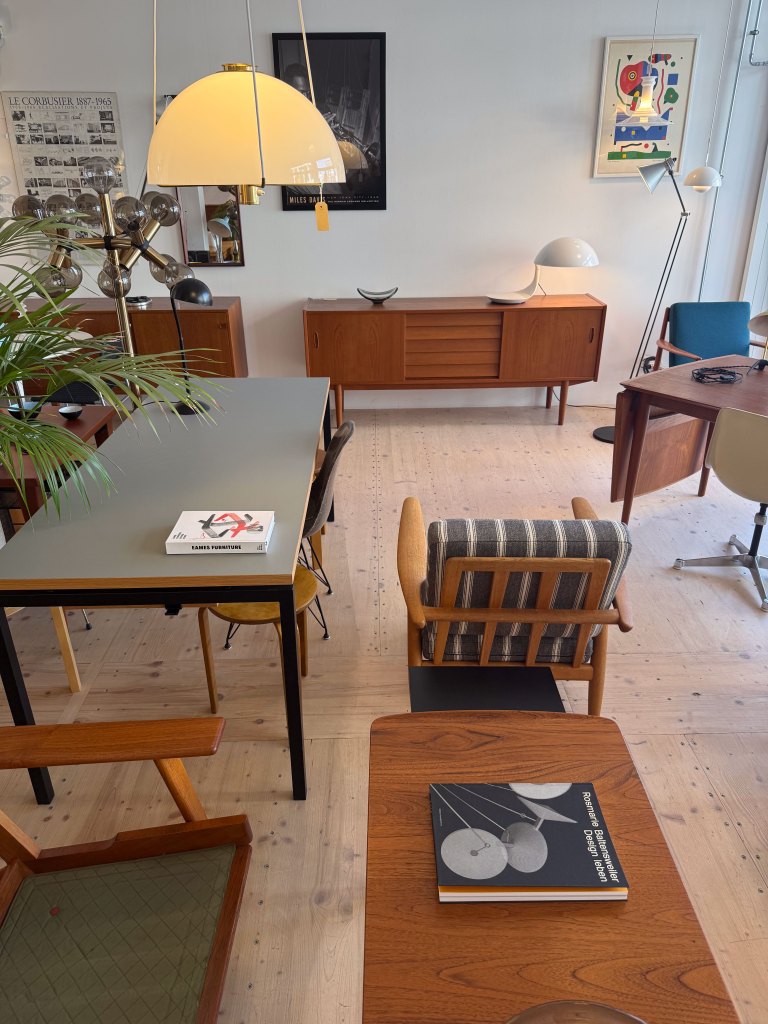 A stylish interior featuring various pieces of mid-century modern furniture, including chairs, tables, and lighting fixtures. A stack of books is visible on a table, alongside a potted plant and decorative items, set against a light wooden floor.