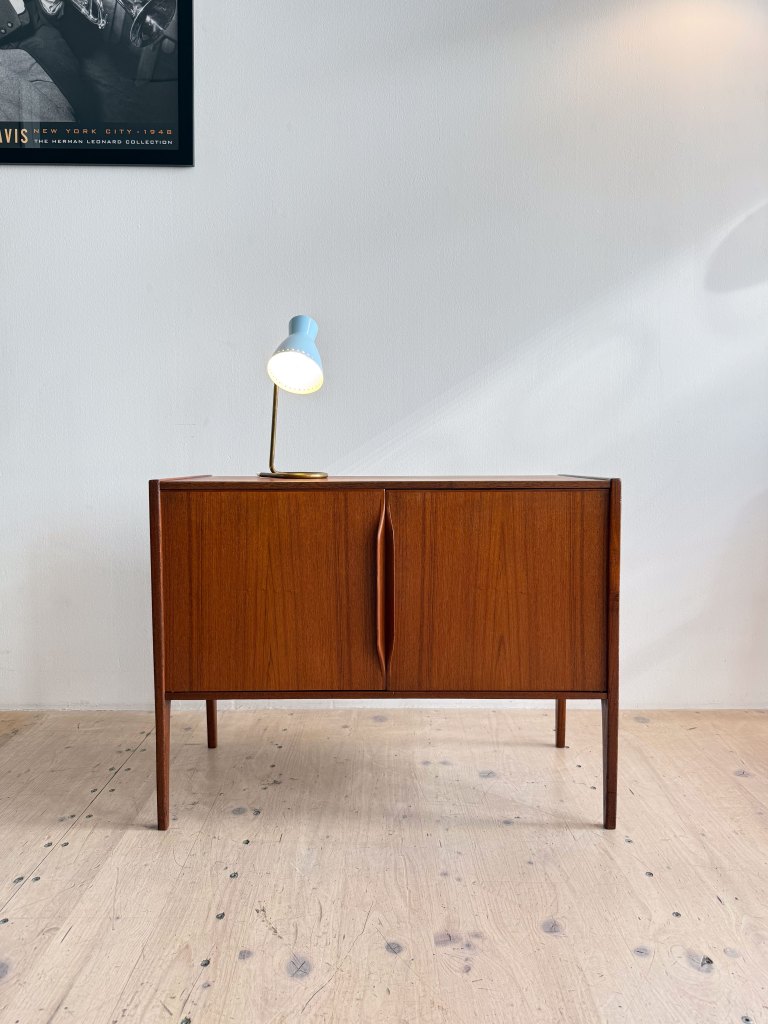 A wooden side cabinet with two doors and a minimalist design, paired with a blue desk lamp on top, set against a light-colored wall.