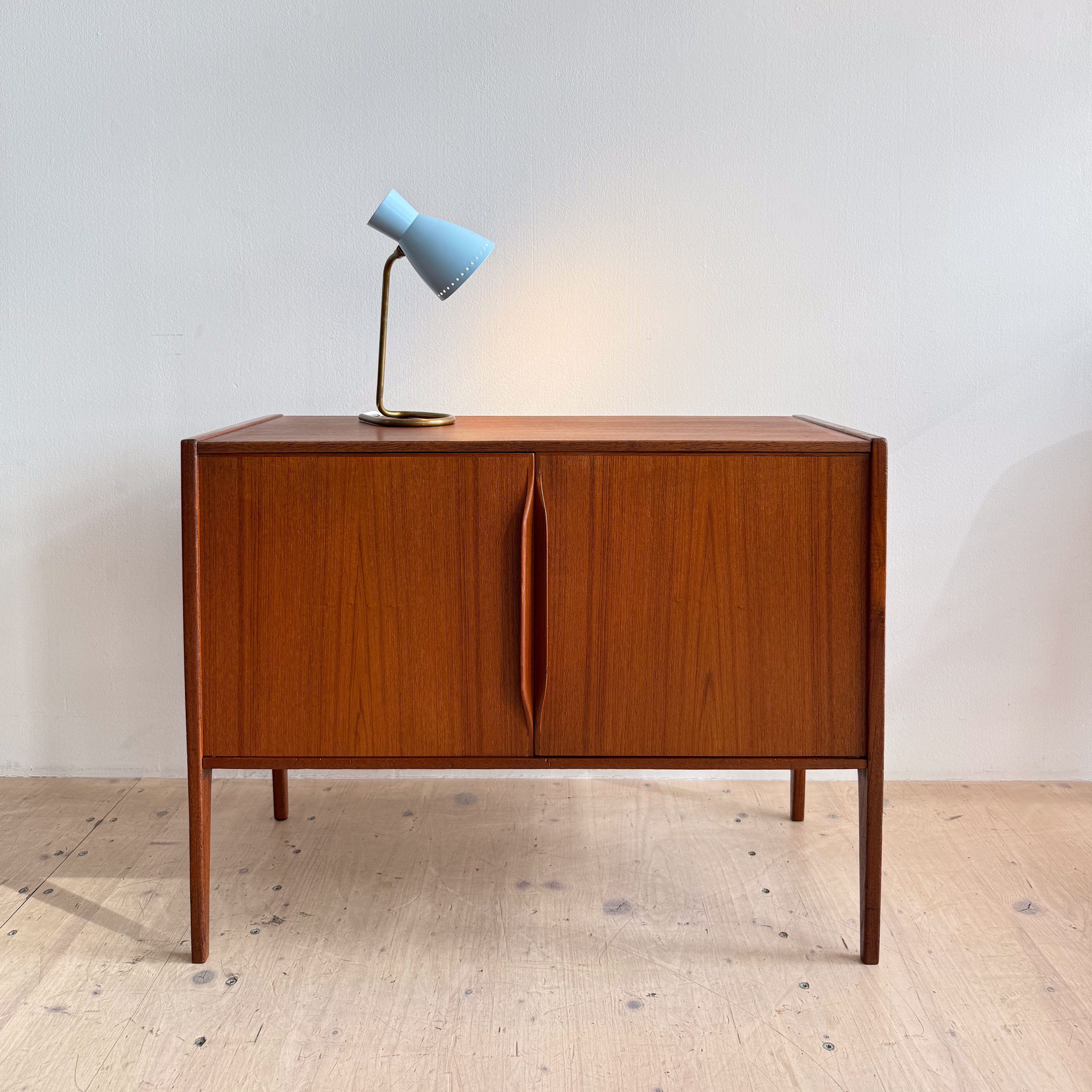 A wooden mid-century modern sideboard with two doors and a sleek design, accompanied by a blue bedside lamp on top.
