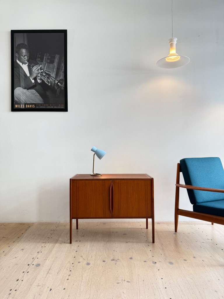 A stylish interior featuring a wooden cabinet, a blue desk lamp, and a modern armchair. A framed black-and-white portrait of a trumpet player hangs on the wall above.