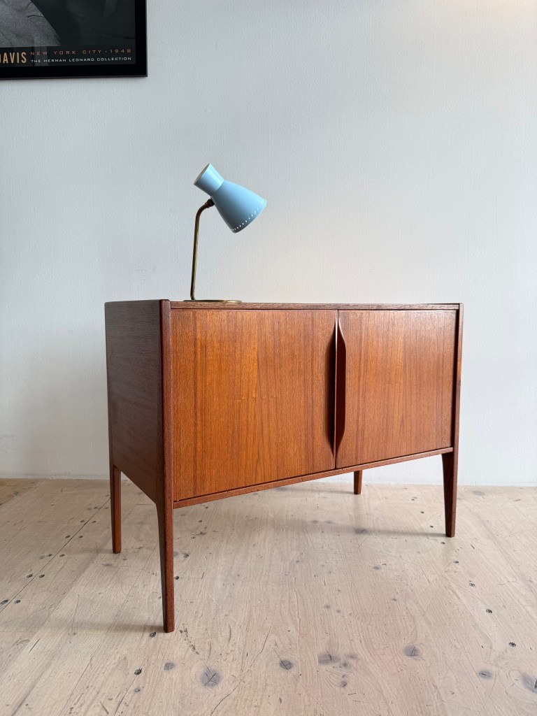 A mid-century wooden sideboard with two cupboard doors and tapered legs, paired with a modern blue desk lamp.