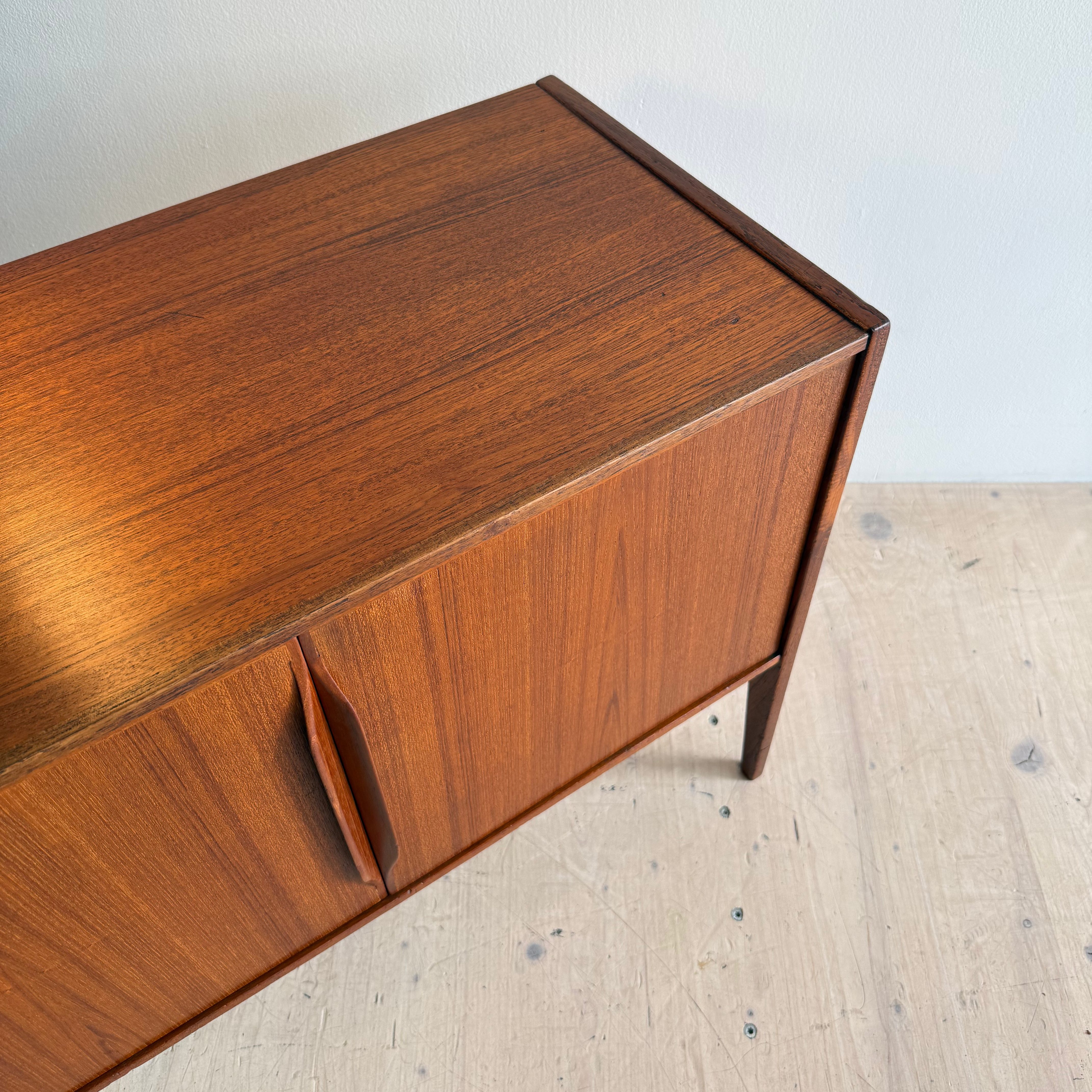 A close-up view of a mid-century modern wooden cabinet featuring a smooth, polished surface and a warm brown finish. The cabinet has two doors with minimalist handles, set against a light-colored floor and wall.