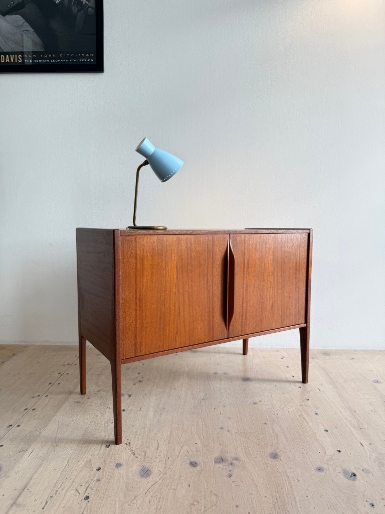 A mid-century modern wooden cabinet with two doors and tapered legs, accompanied by a blue desk lamp on top.