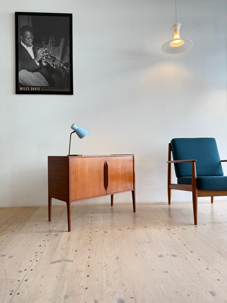 A modern living space featuring a wooden cabinet, a blue table lamp, and a teal upholstered armchair. A black and white framed poster of a musician hangs on the wall above the cabinet.