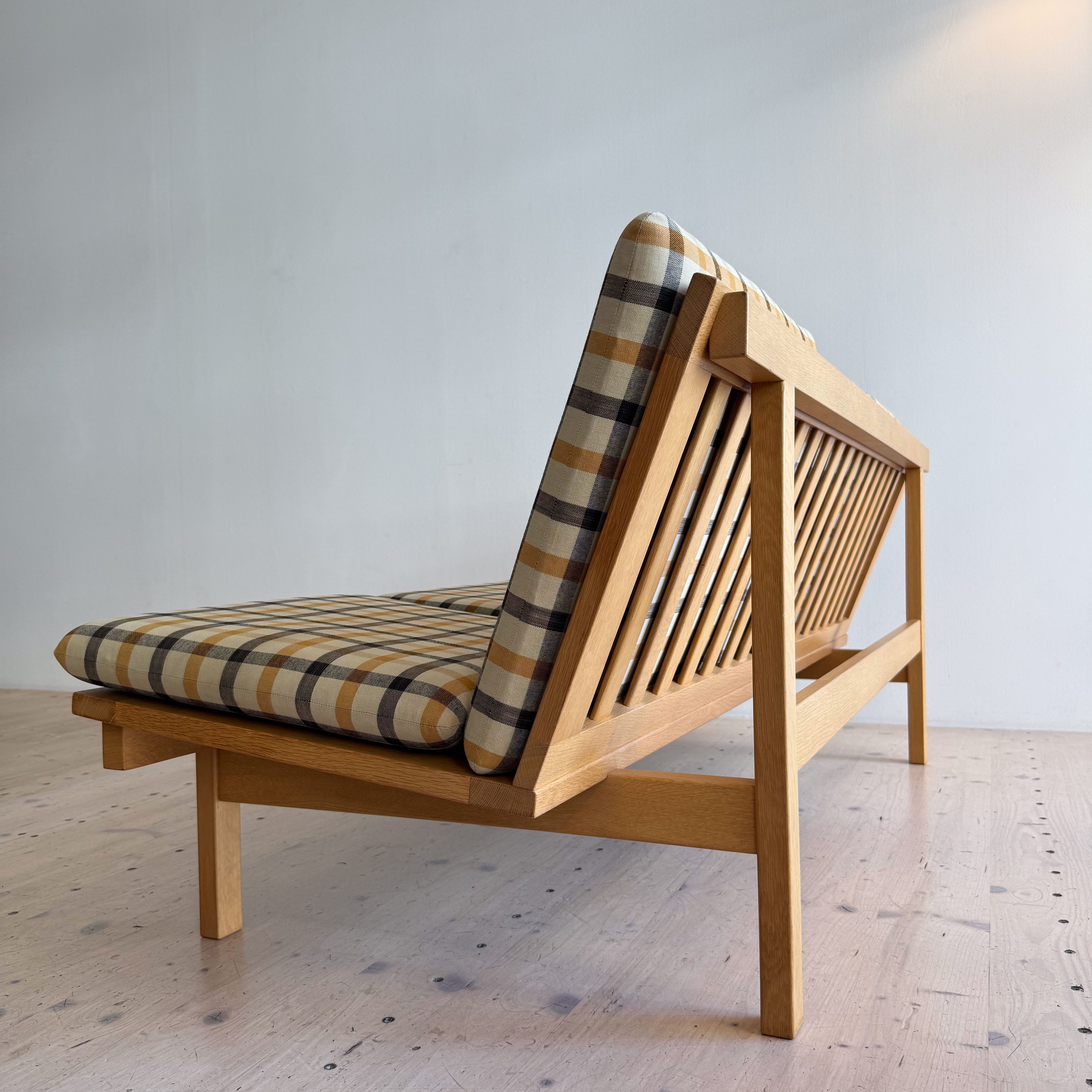 A mid-century modern wooden bench with a slatted back and a plaid fabric cushion, set against a minimalistic white wall.