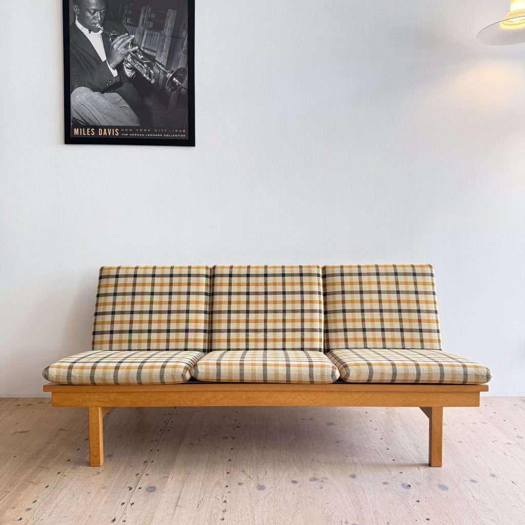A vintage-style sofa with plaid upholstery in yellow, brown, and green tones, placed against a white wall. Above it hangs a black and white poster of a musician playing the trumpet.