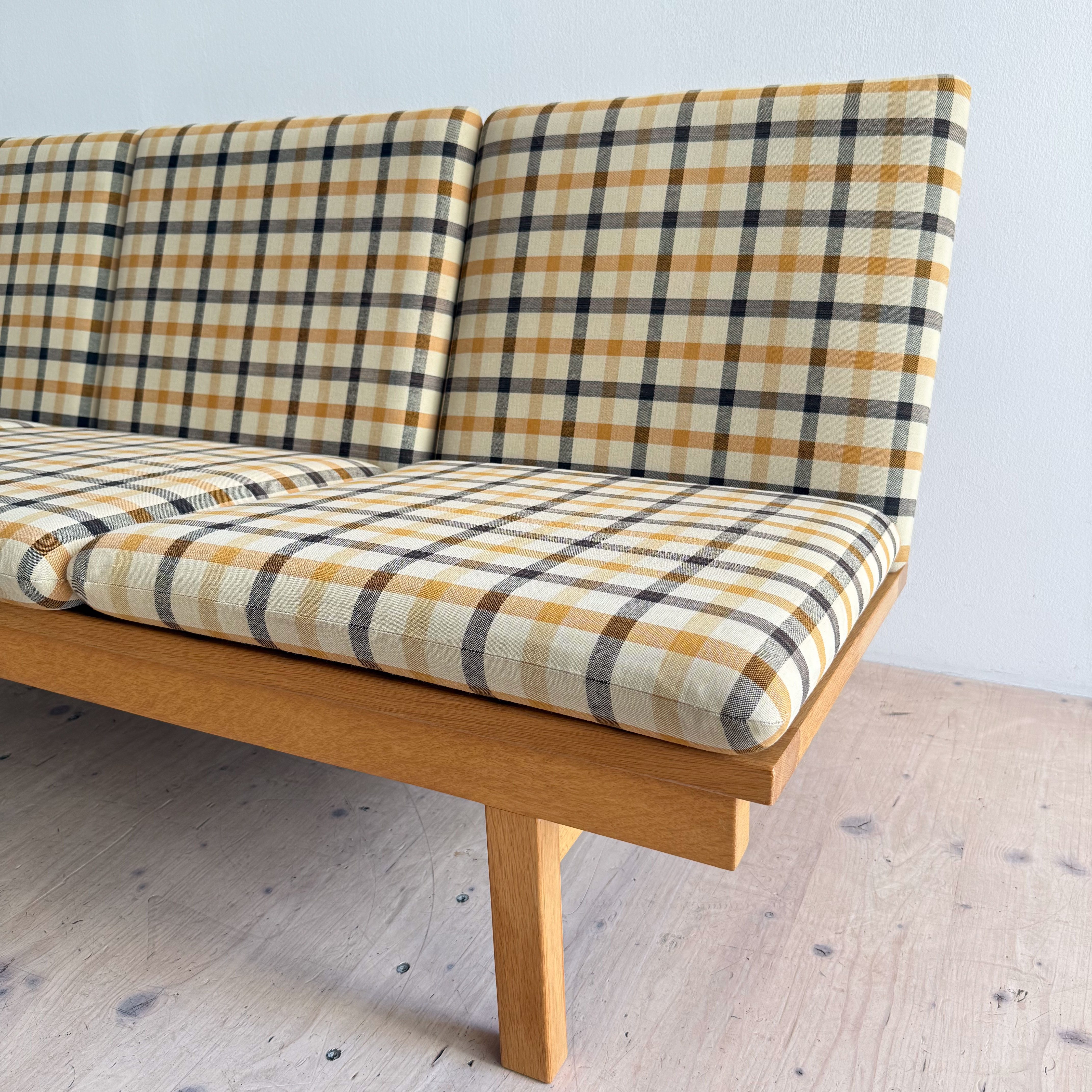 A modern wooden bench with a padded seat and backrest featuring a yellow, black, and white plaid pattern, set against a light wall.