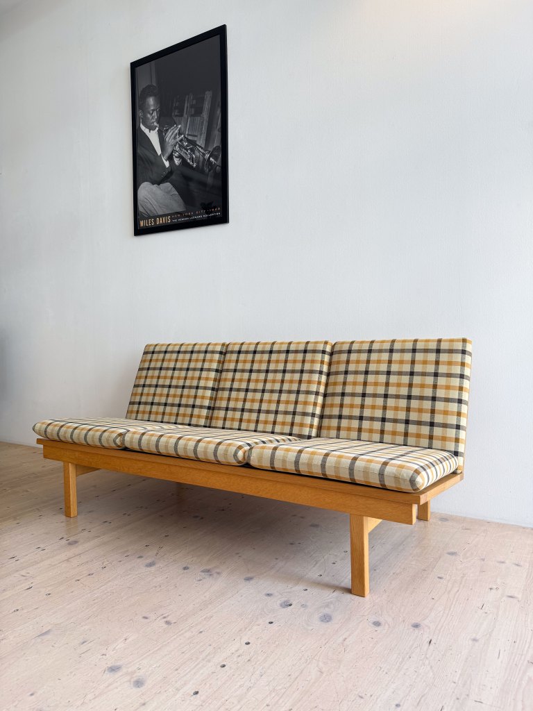 A plaid patterned sofa with wooden legs, positioned against a white wall featuring a framed black and white poster of a musician.