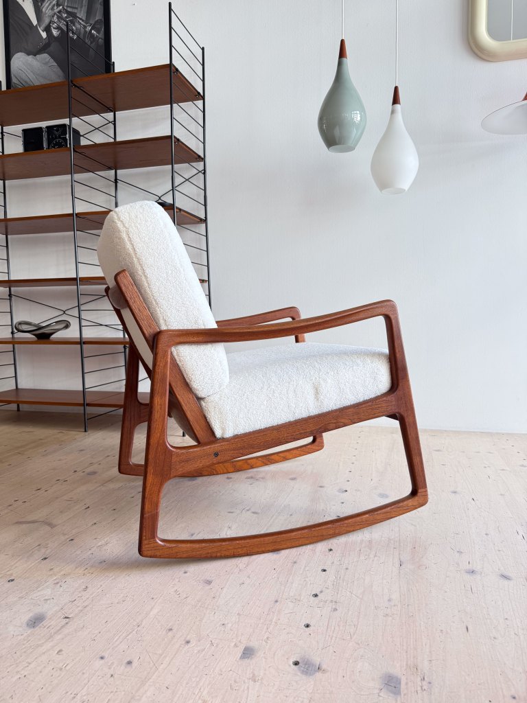 A modern wooden rocking chair with a light-colored upholstered seat, positioned in a bright interior space featuring a minimalist shelving unit and decorative pendant lights.