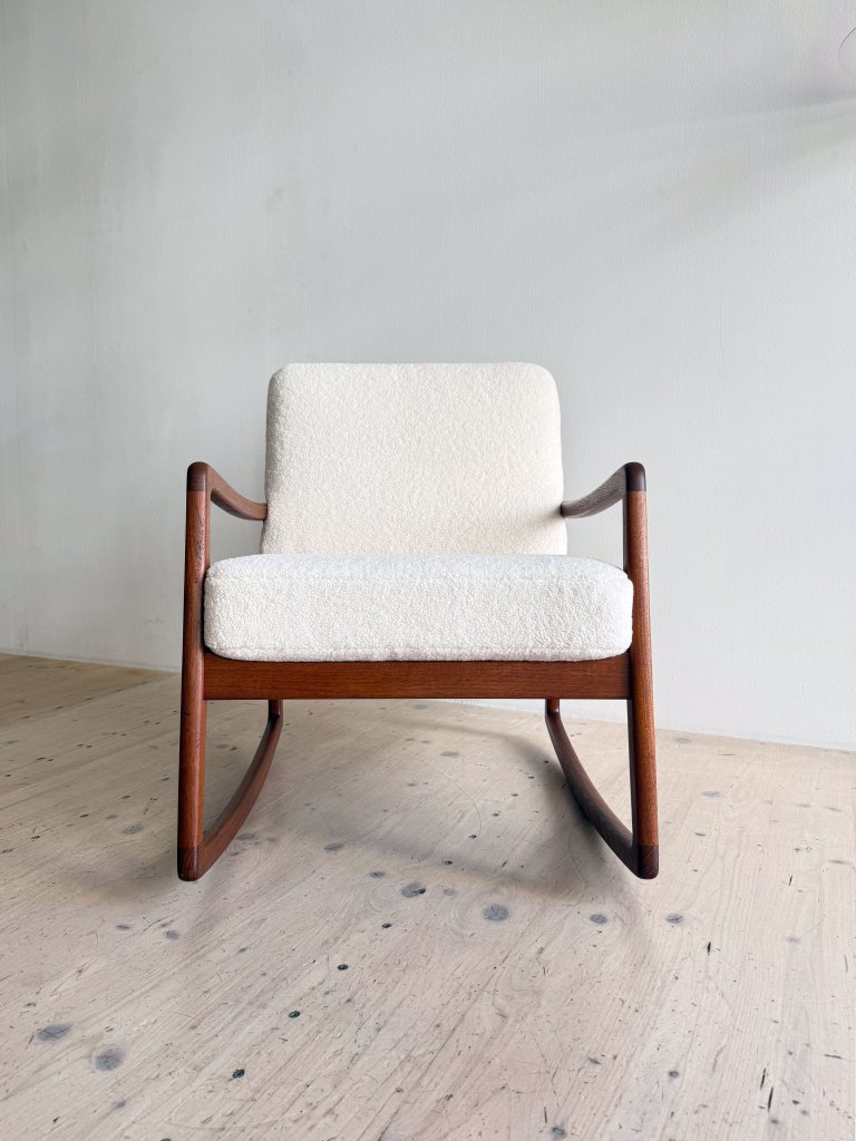 A modern rocking chair with a wooden frame and a soft, cream-colored upholstered seat, positioned against a light grey wall.