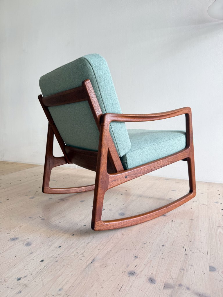 A mid-century modern rocking chair with a wooden frame and soft green upholstery, set against a light-colored wall and wooden floor.