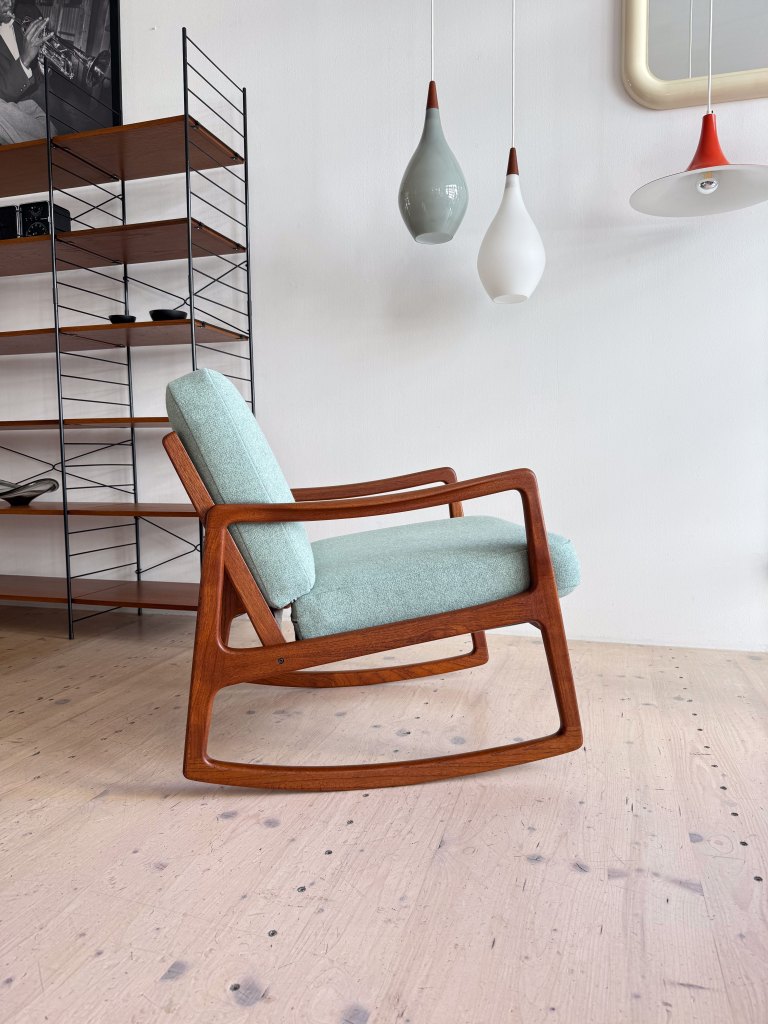 A wooden rocking chair with a light blue cushion, positioned in a minimalist interior space with a wooden floor and decorative hanging lights.