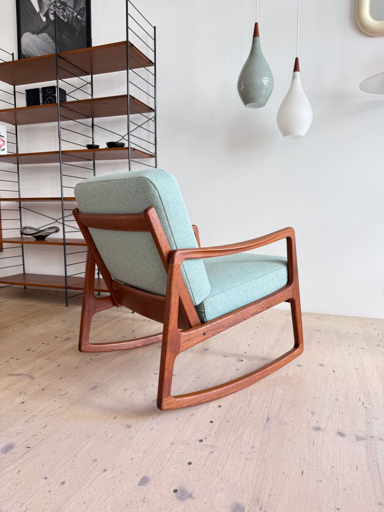 A modern rocking chair with a light blue cushion and wooden frame, positioned next to a metal shelf displaying various decorative items. Two pendant lights are hanging above.