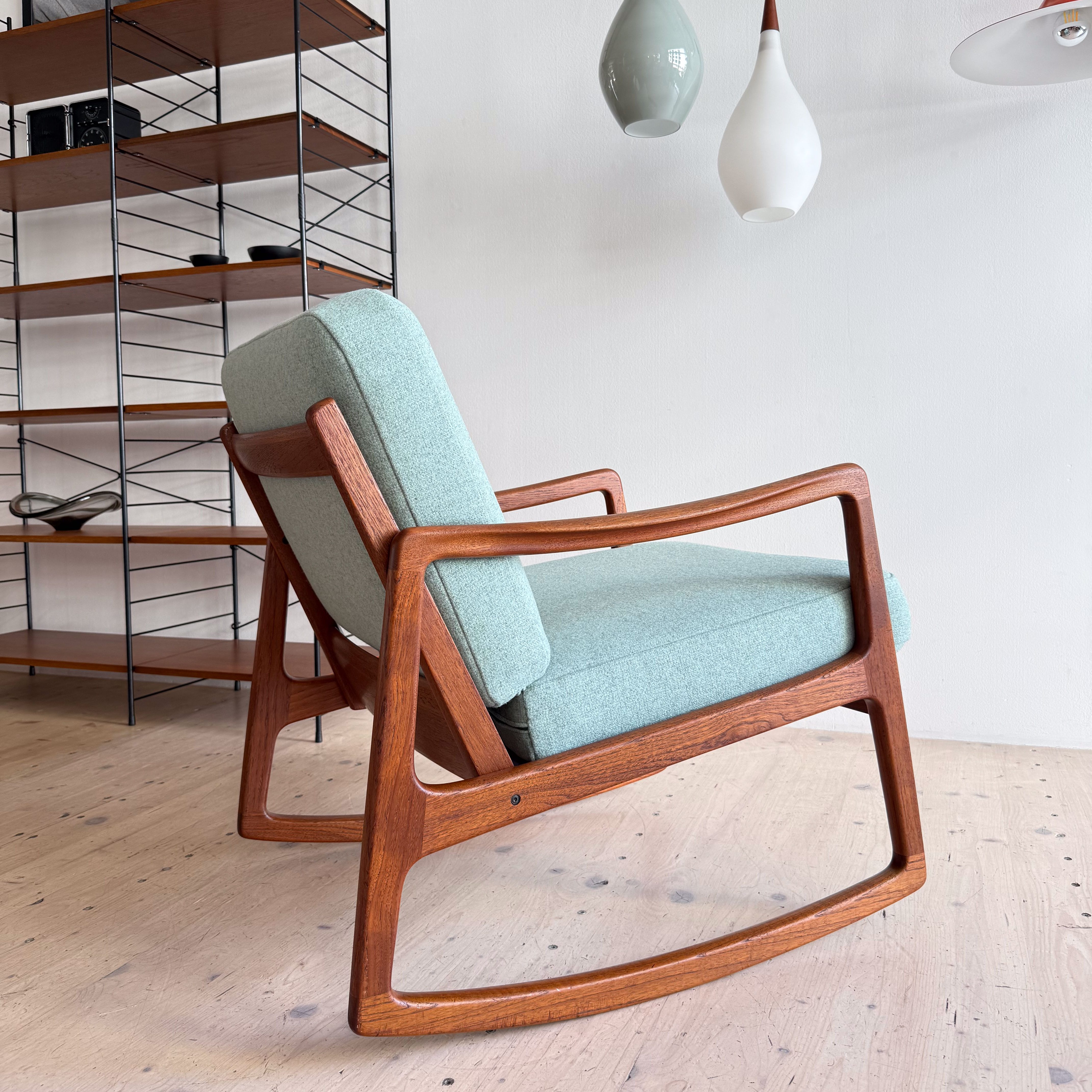 A mid-century modern rocking chair with a light blue upholstered seat and wooden frame, positioned in a minimalist living space featuring a wooden shelf and pendant lighting.